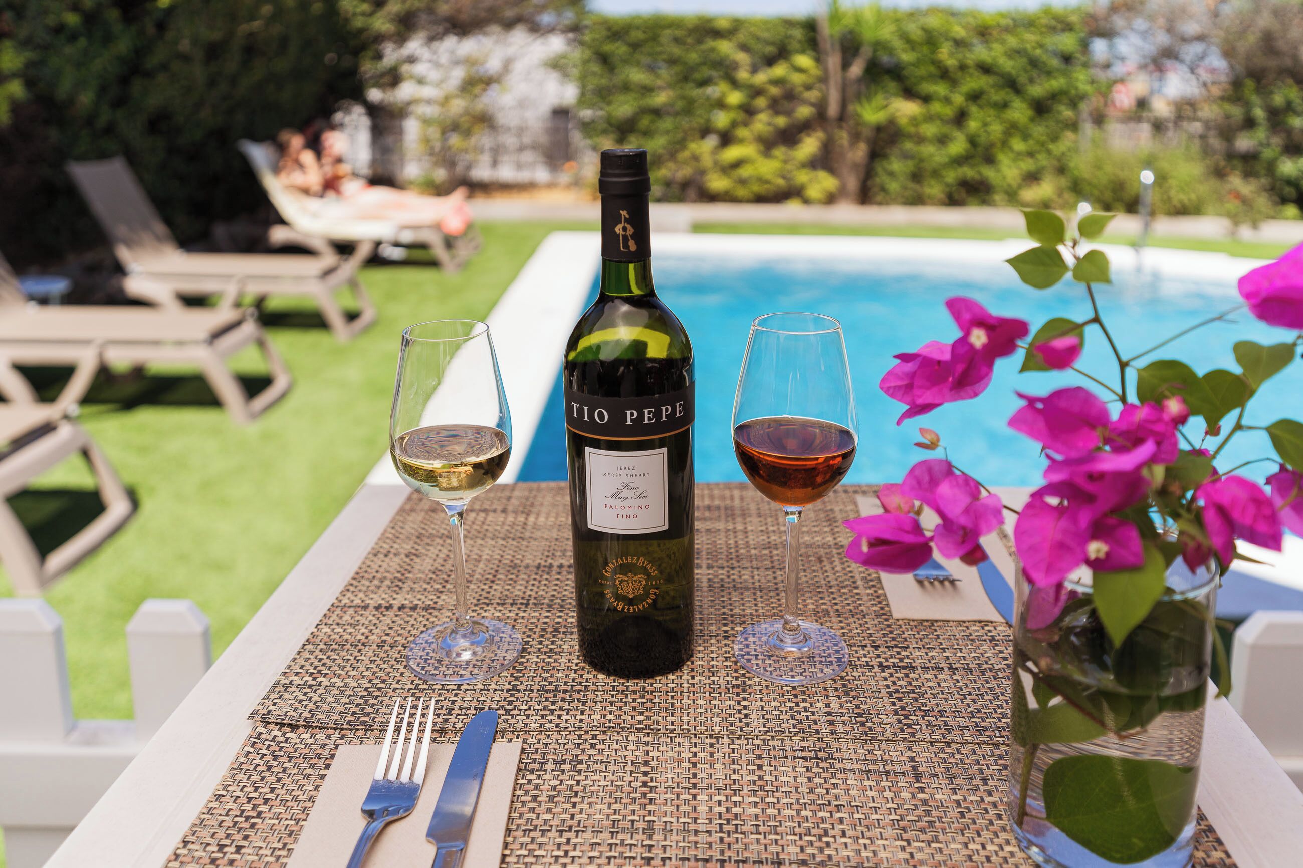 Una botella de vino Tio Pepe, dos copas de vino, un tenedor y un cuchillo sobre una mesa con un mantel tejido, cerca de una piscina con sillas de descanso y un jarrón con flores.