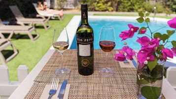 Una botella de vino Tio Pepe, dos copas de vino, un tenedor y un cuchillo sobre una mesa con un mantel tejido, cerca de una piscina con sillas de descanso y un jarrón con flores.