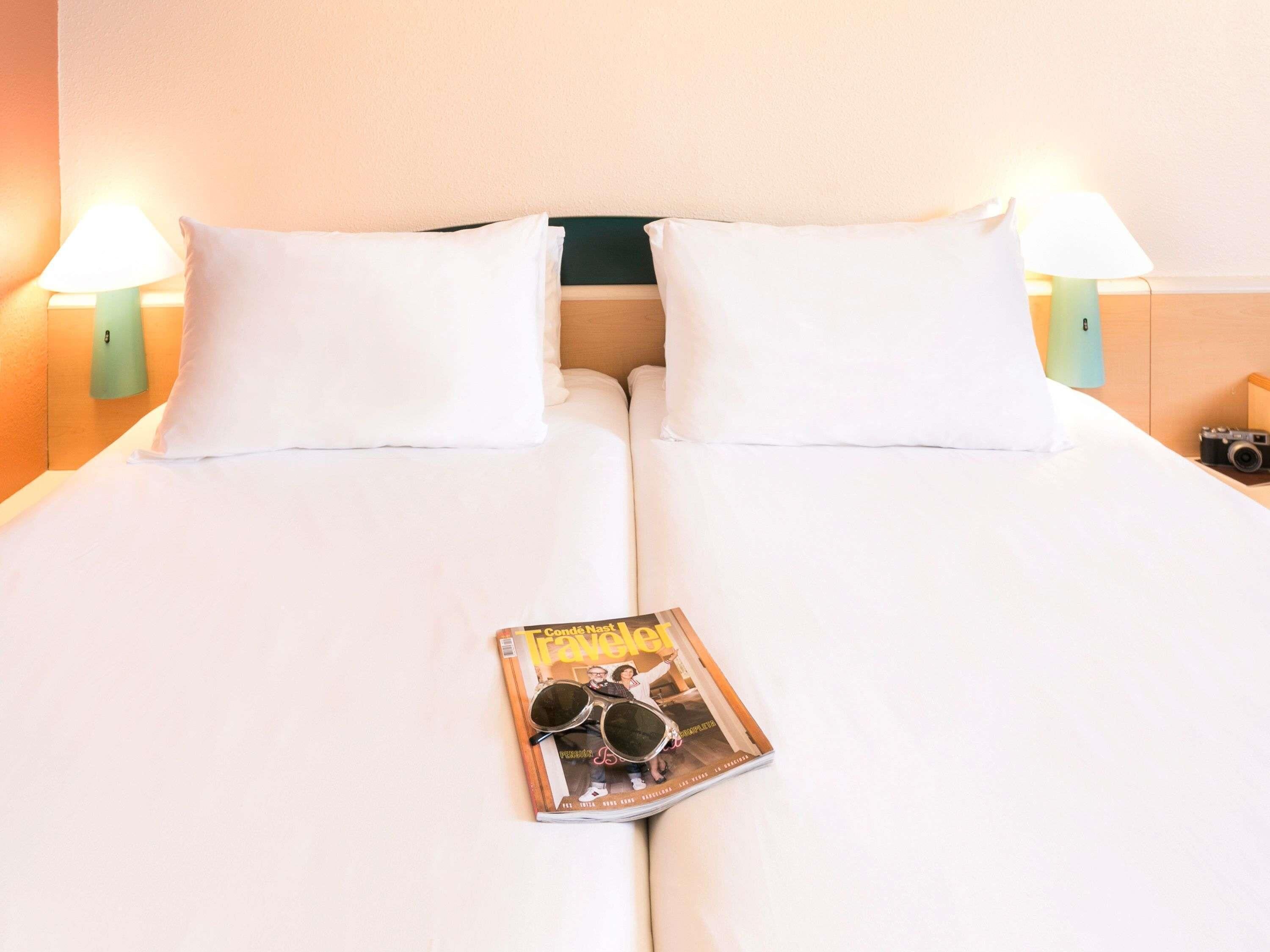A neatly made bed with white linens and two pillows, a magazine with sunglasses on it, and two wall-mounted lamps on either side.