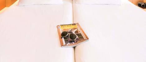 A neatly made bed with white linens and two pillows, a magazine with sunglasses on it, and two wall-mounted lamps on either side.