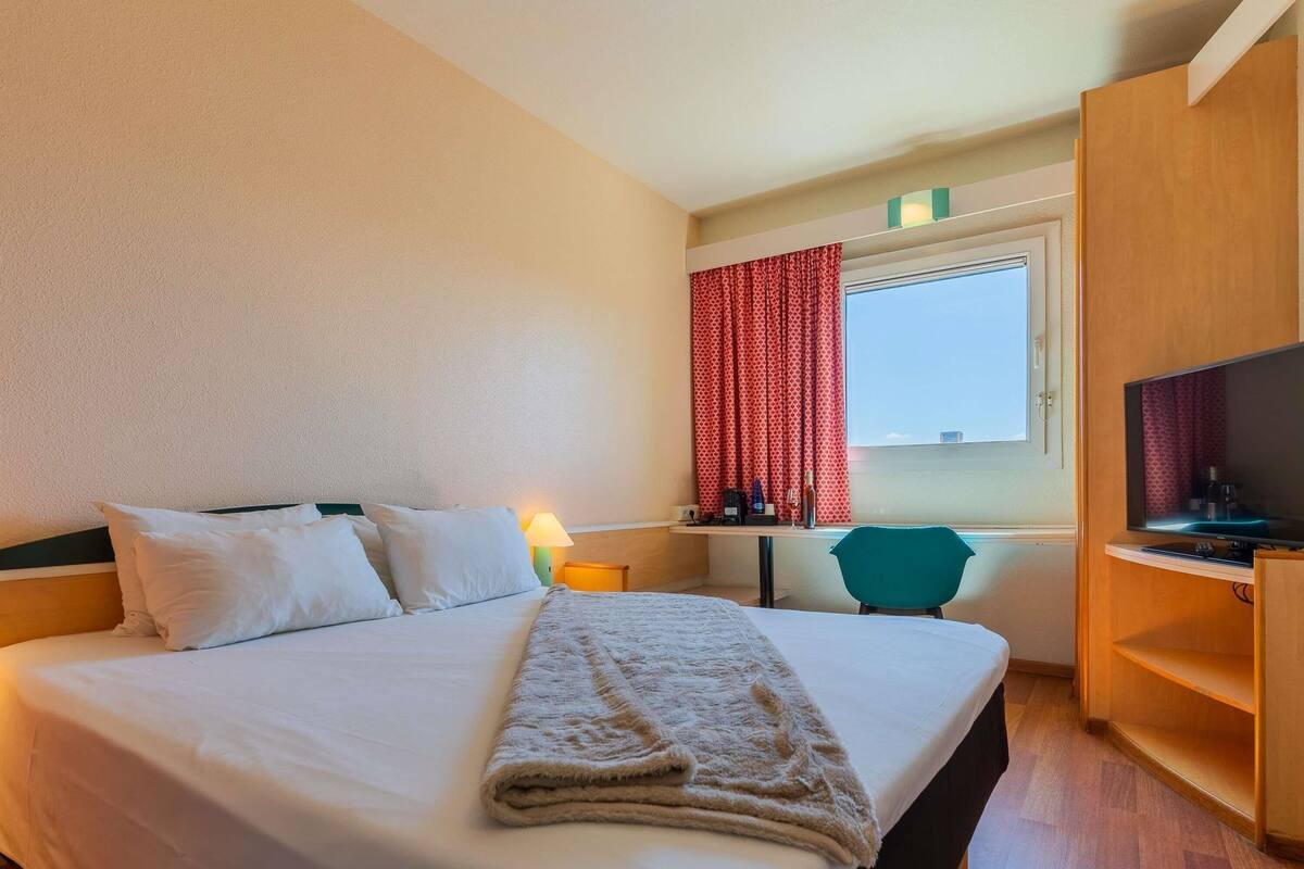 Une chambre d’hôtel équipée d’un lit, d’un bureau, d’une chaise, d’une télévision et d’une fenêtre avec des rideaux.