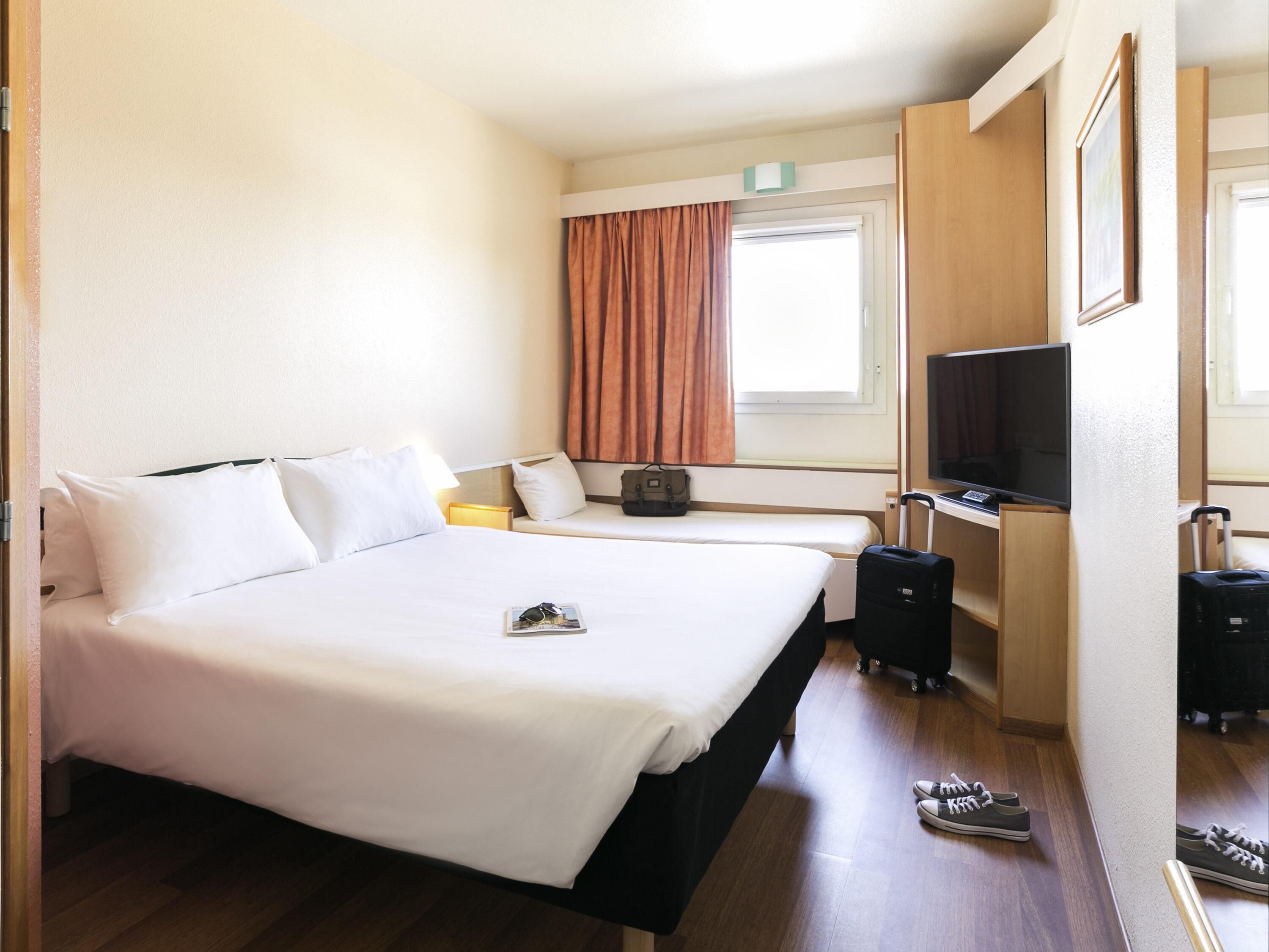 Une chambre d’hôtel avec un grand lit, un bureau avec une télévision, une valise et une fenêtre avec des rideaux.