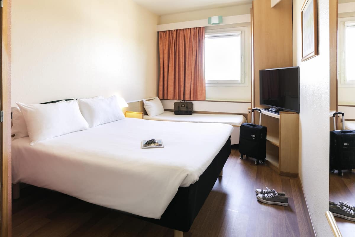 A hotel room with a large bed, a desk with a television, a suitcase, and a window with curtains.