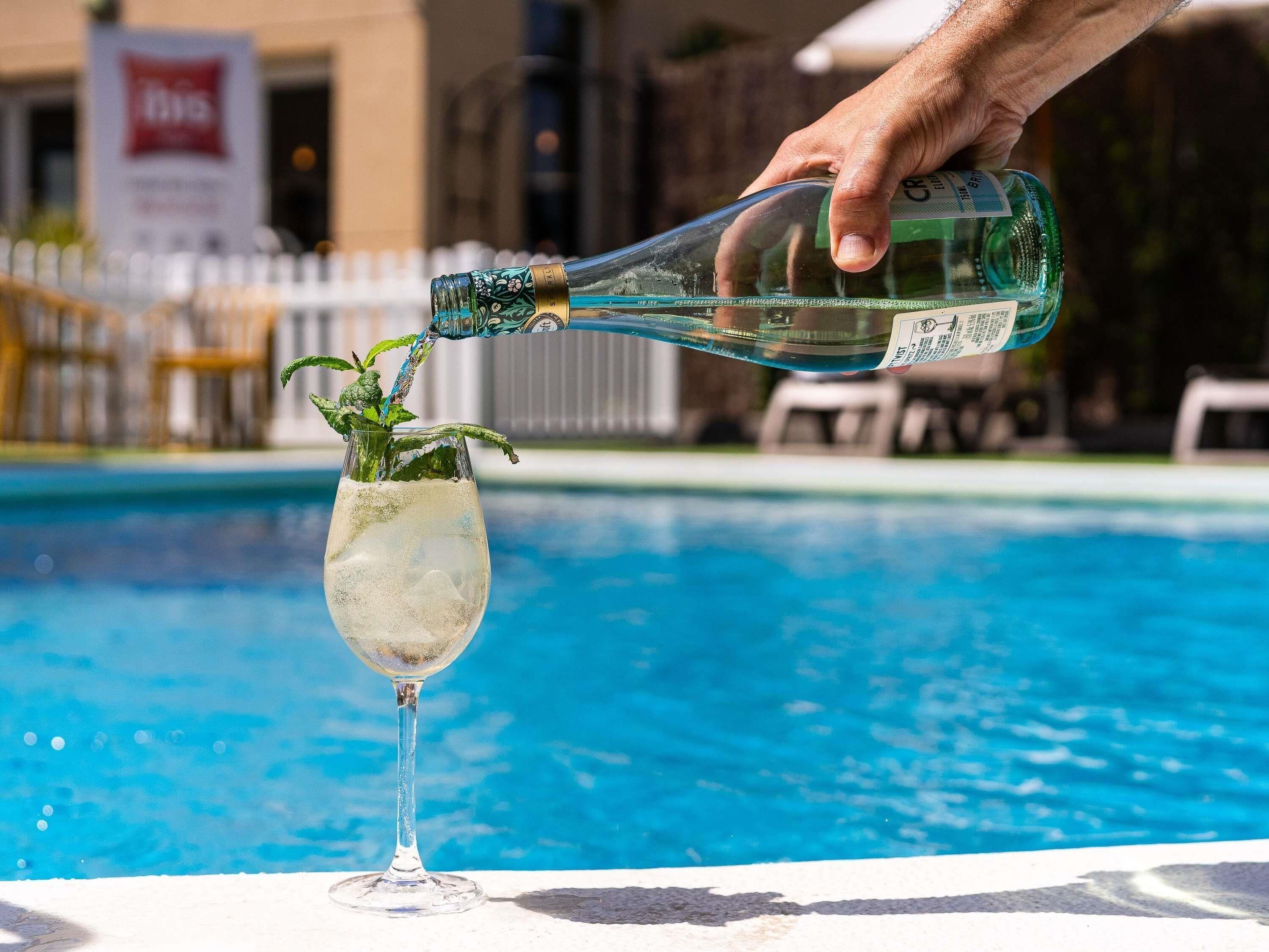 Une main verse une boisson d’une bouteille verte dans un verre, qui est décoré d’une feuille de menthe, le tout près d’une piscine.