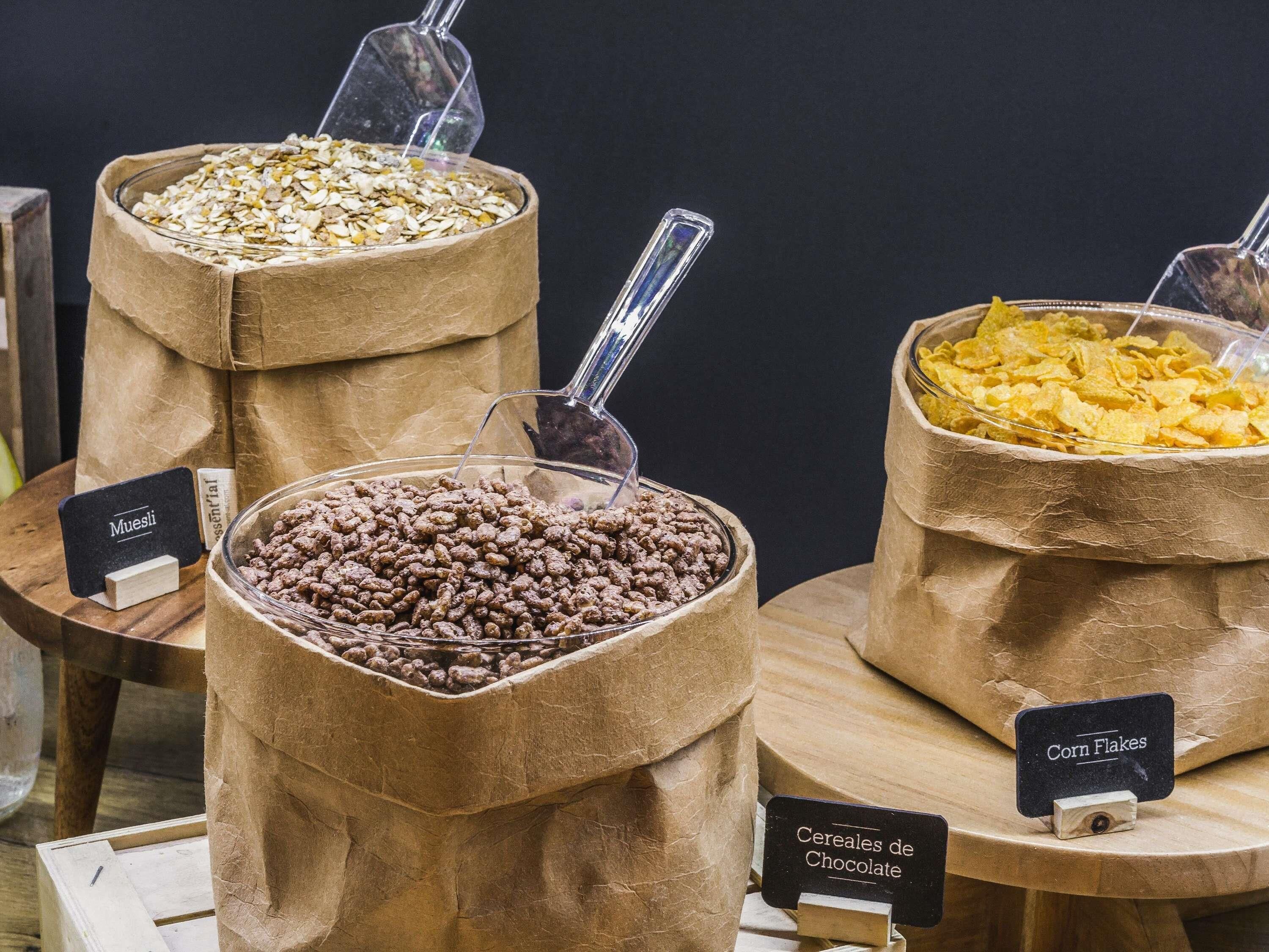 Tres bolsas de papel con etiquetas para distintos alimentos, un soporte de madera y un fondo oscuro.