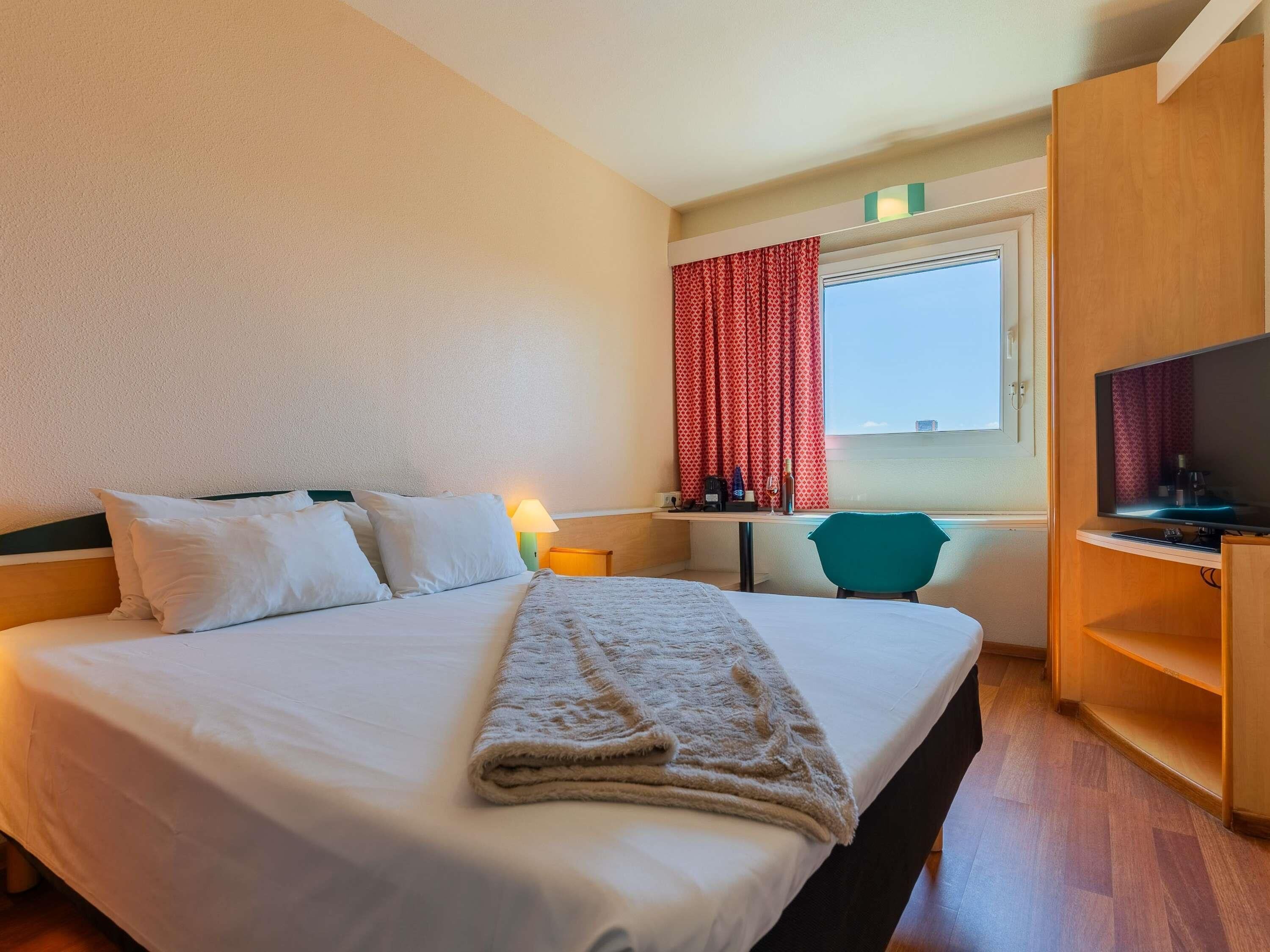 Une chambre d’hôtel avec un lit, un bureau et une chaise, une télévision et une fenêtre avec des rideaux.