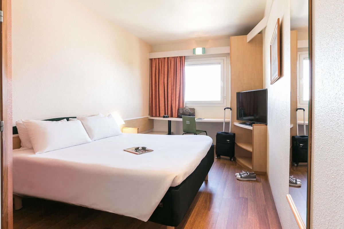 Une chambre d’hôtel avec un lit, un bureau, une télévision et une fenêtre avec des rideaux.