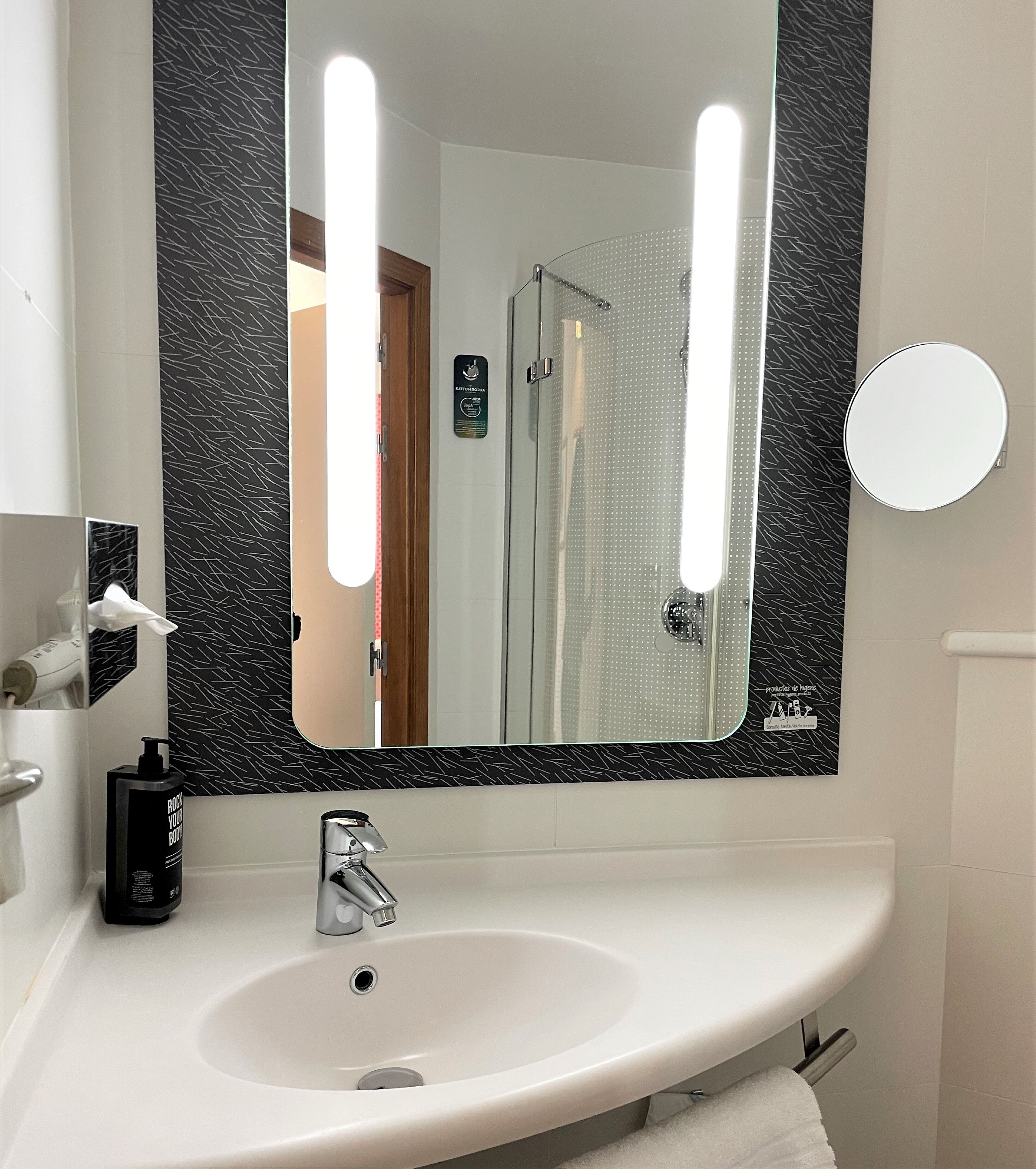 Un baño con lavabo blanco, espejo con iluminación, toalla y puerta de ducha.