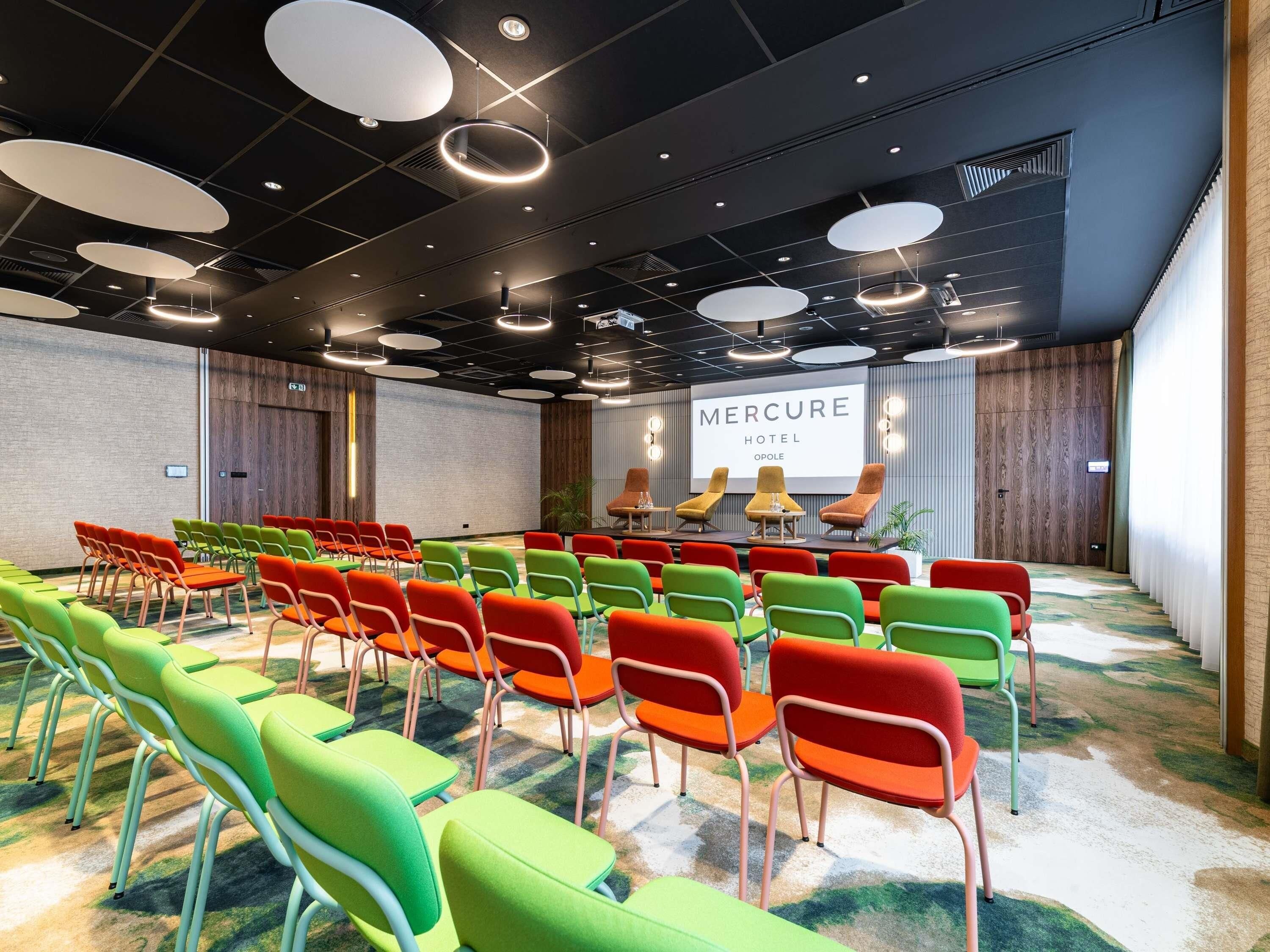 A conference room with rows of colorful chairs, a stage with a screen displaying 'MERCURE HOTEL OPOLE,' and a modern ceiling design.