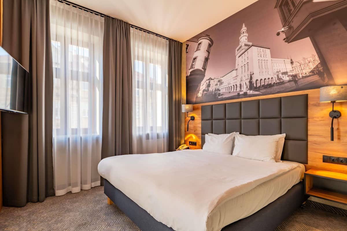 Habitación de hotel con una cama grande, un televisor, un cuadro de un edificio y un teléfono en la mesita de noche.
