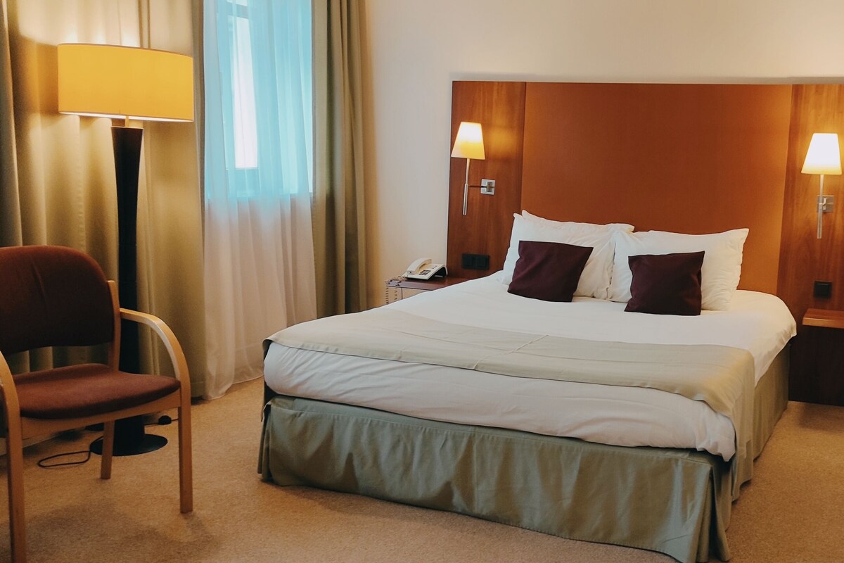 A hotel room with a large bed, two bedside lamps, a chair, and a desk with a lamp.