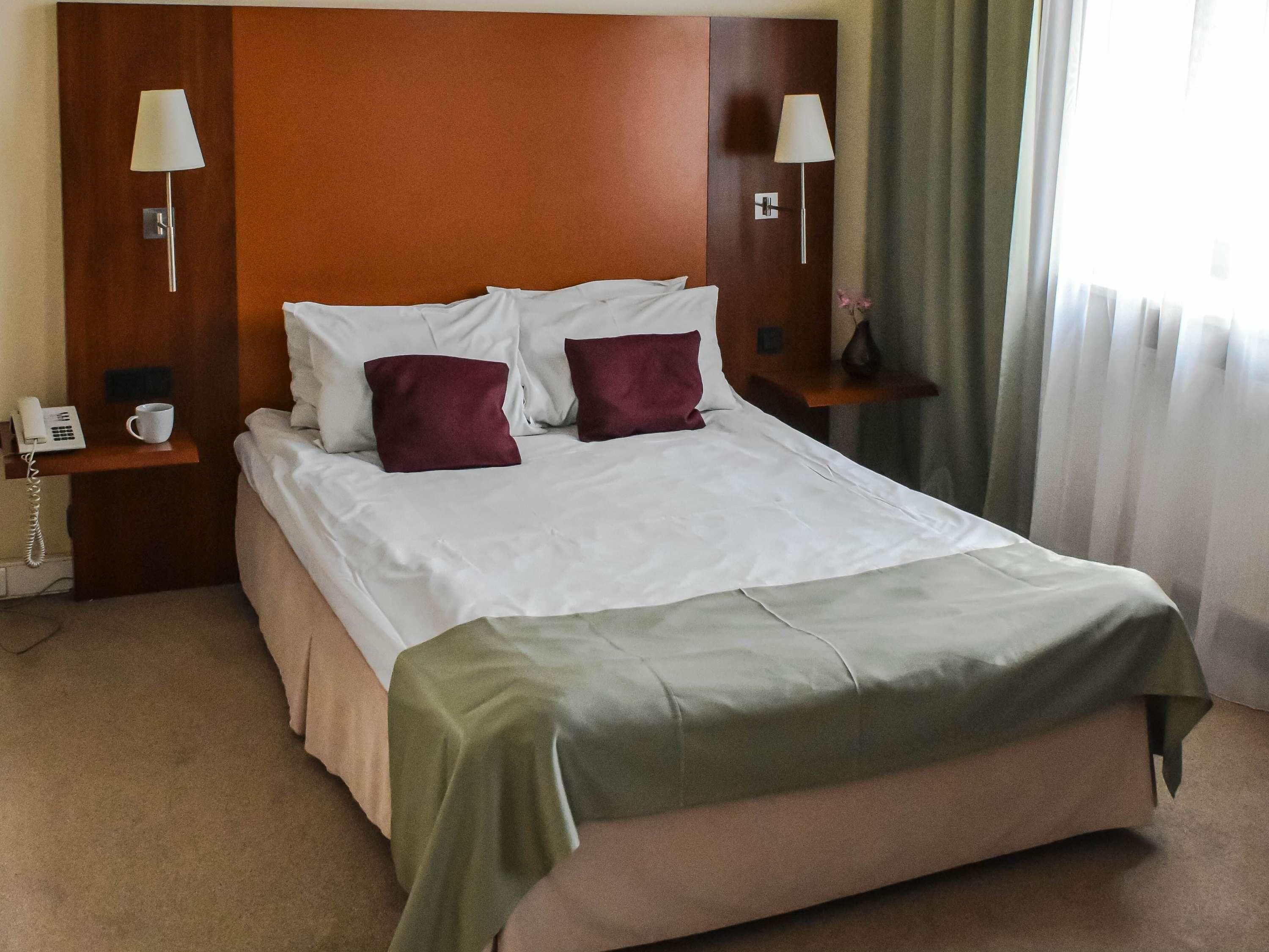 A hotel room with a large bed, two bedside tables with lamps, a telephone, and a cup on one table.