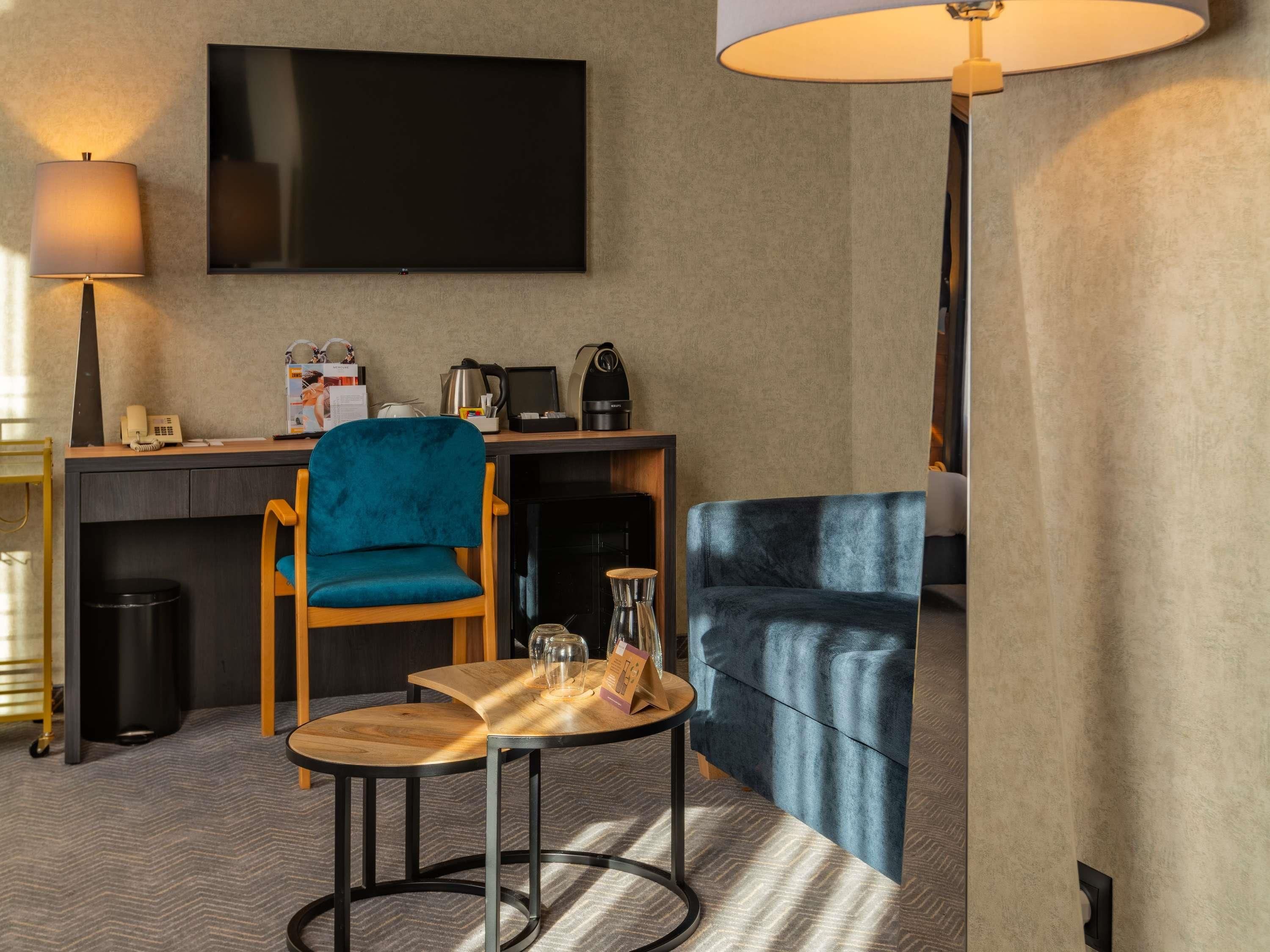Habitación de hotel con televisión de pantalla plana, escritorio con cafetera, una silla azul, una mesa de madera y un sofá.
