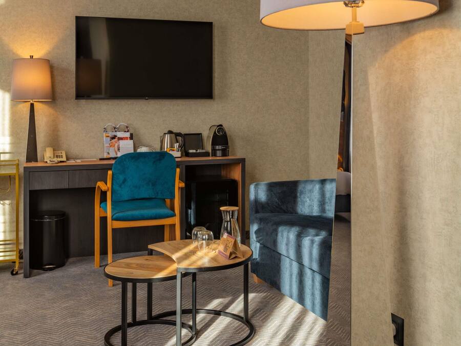 Habitación de hotel con televisión de pantalla plana, escritorio con cafetera, una silla azul, una mesa de madera y un sofá.