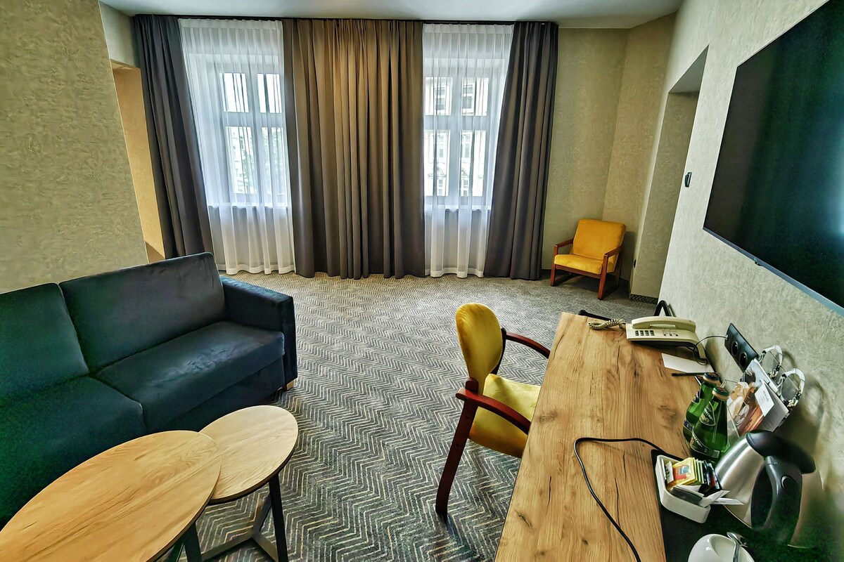 A hotel room with a sofa, a yellow armchair, a wooden table, and a TV mounted on the wall.