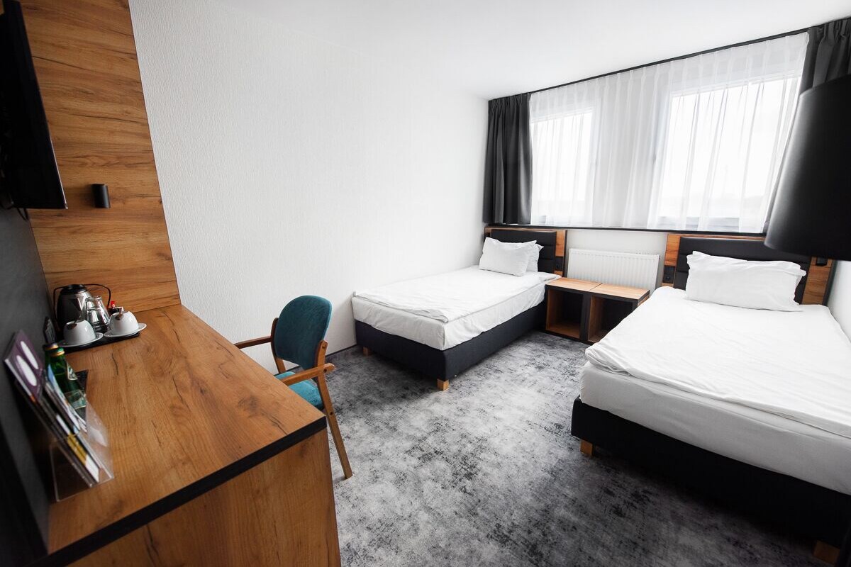 A hotel room with two beds, a wooden desk, a chair, a coffee maker, and a large window with curtains.