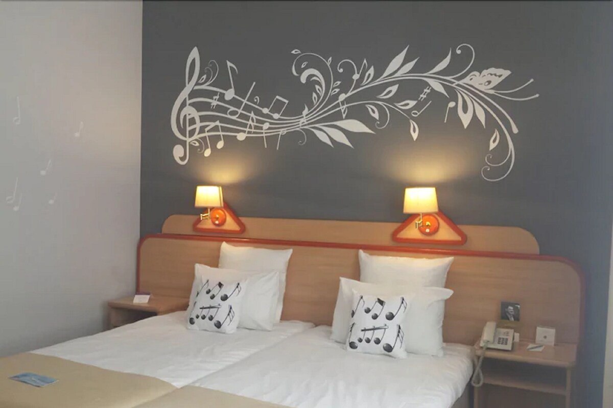 A hotel room with a bed, two bedside lamps, a nightstand with a phone, and a decorative wall with musical notes and leaves.