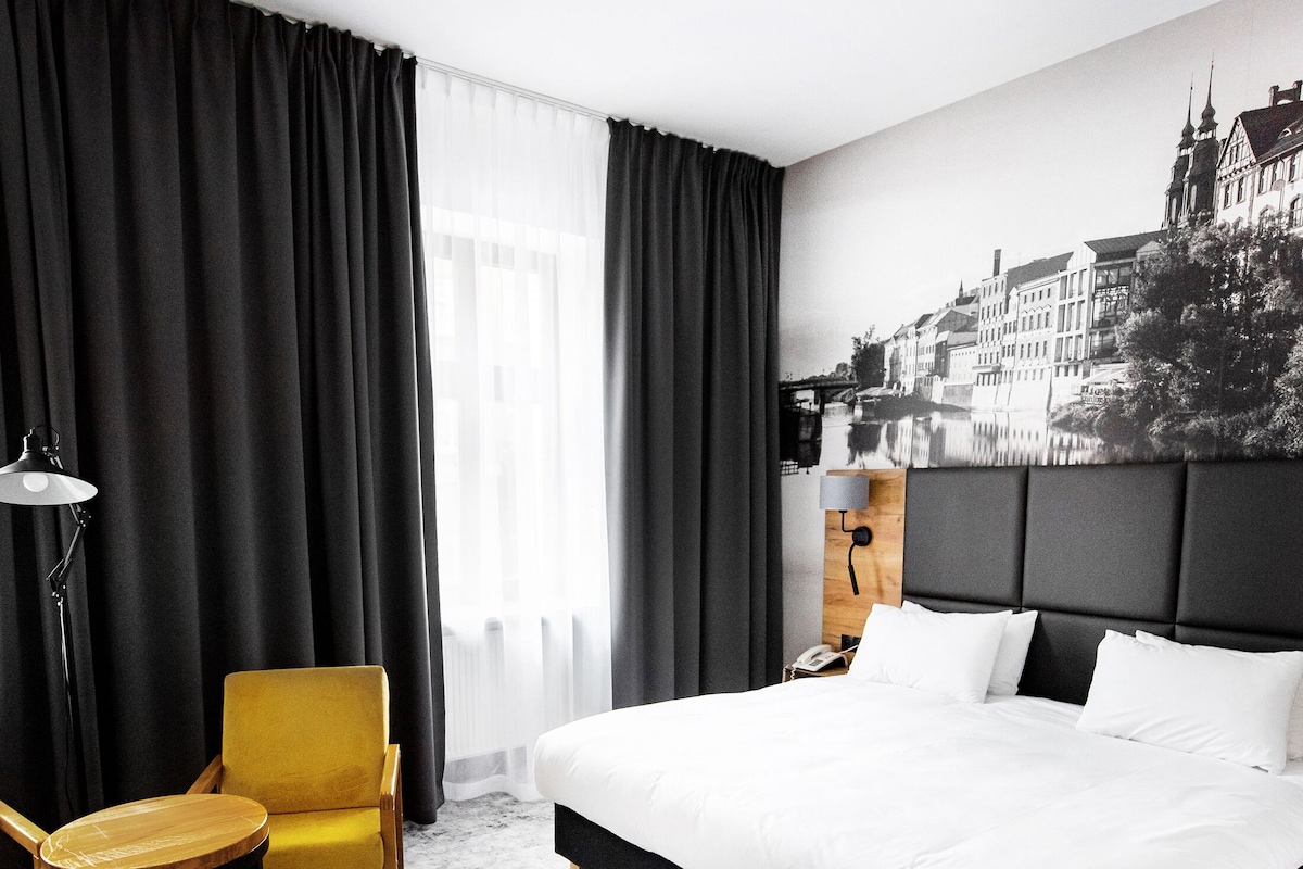 A hotel room with a large bed, a chair, a table, and a view of a cityscape through sheer curtains.