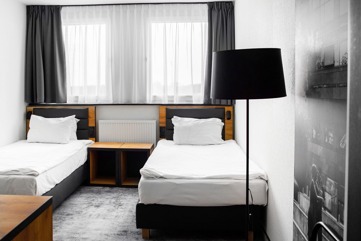 A hotel room with two single beds, a wooden nightstand, a desk, and a large window with sheer curtains.