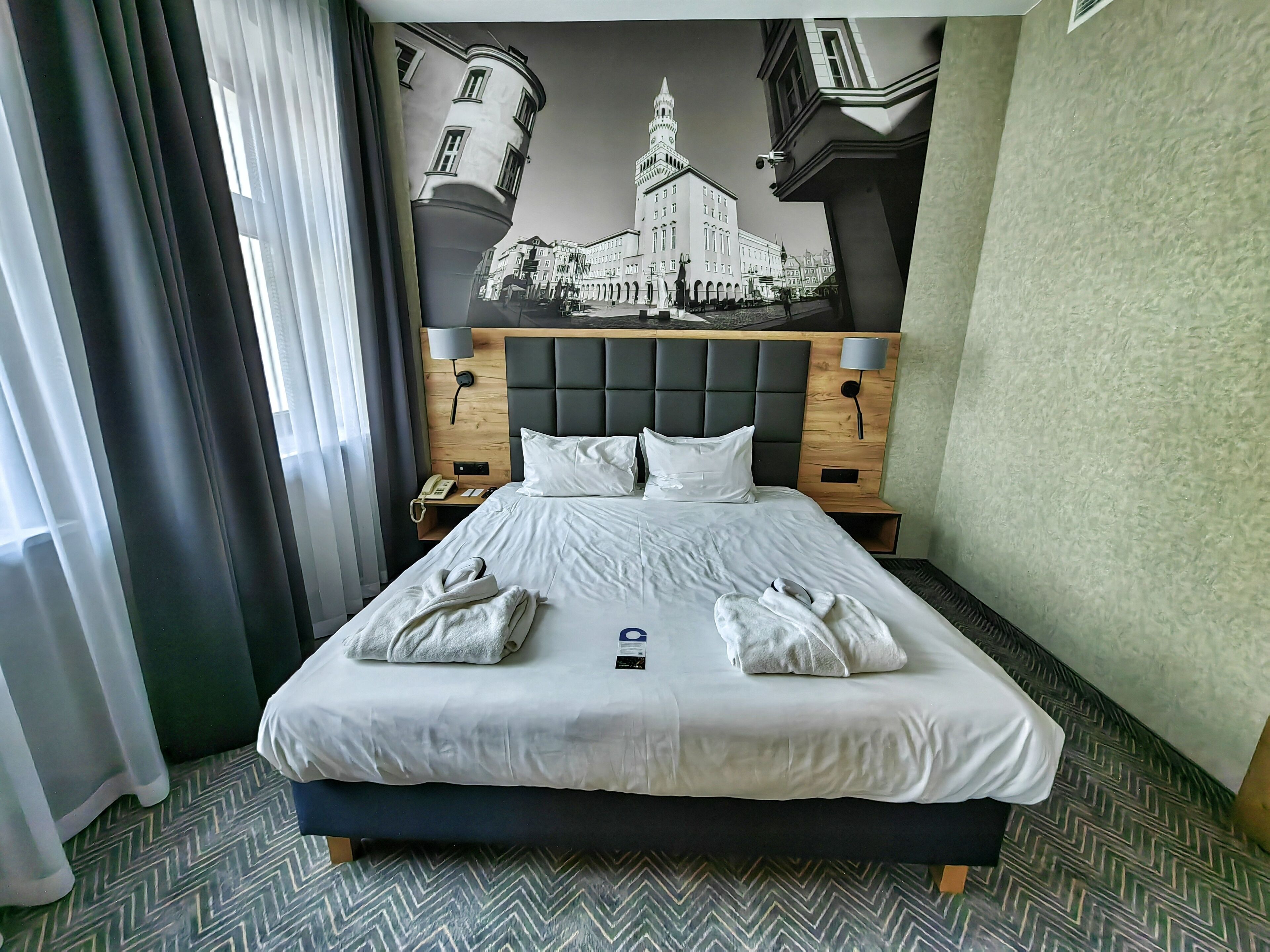 Quarto de hotel com uma cama grande, cabeceira com relógio, mesa de cabeceira com telefone e um mural na parede retratando um edifício.