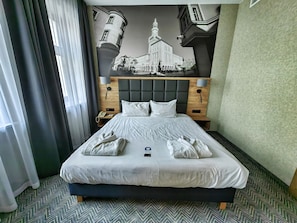 A hotel room with a large bed, a headboard with a clock, a nightstand with a phone, and a wall mural of a building.
