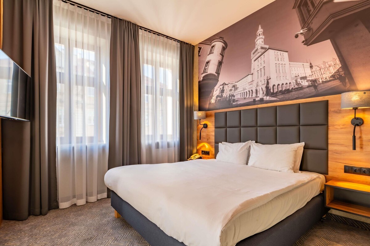 A hotel room with a large bed, a television, a painting of a building, and a telephone on the nightstand.