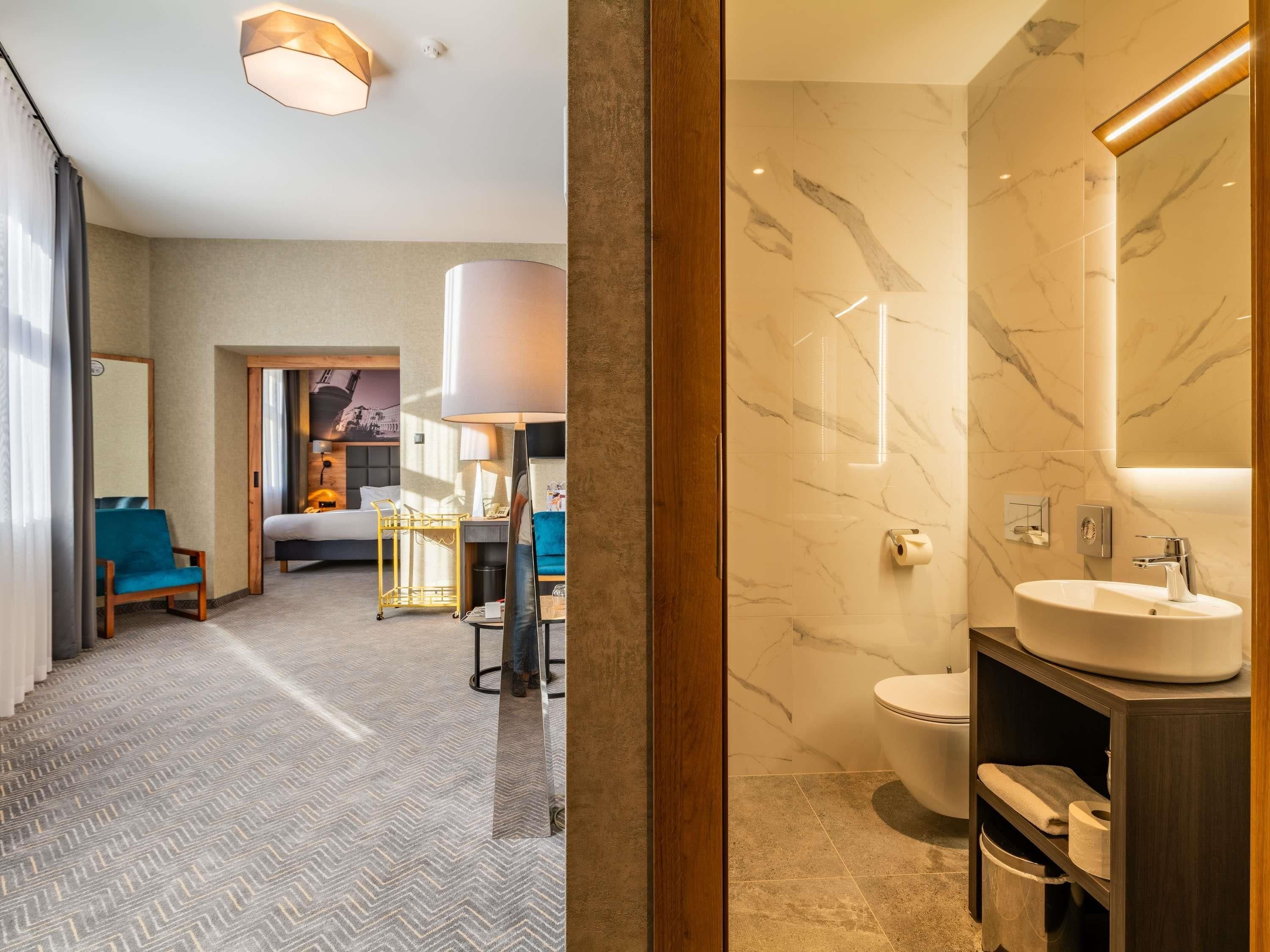 A hotel room with a bed, a desk, a chair, a bathroom with a sink and mirror, and a hallway with a patterned carpet.