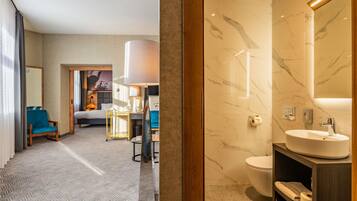 A hotel room with a bed, a desk, a chair, a bathroom with a sink and mirror, and a hallway with a patterned carpet.