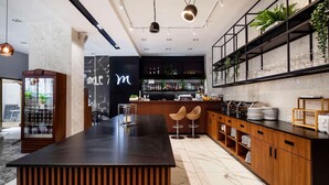 A modern bar area with a marble floor, wooden bar counter, and bar stools. There are shelves with glasses and bottles, and a large 'm' logo on the wall.