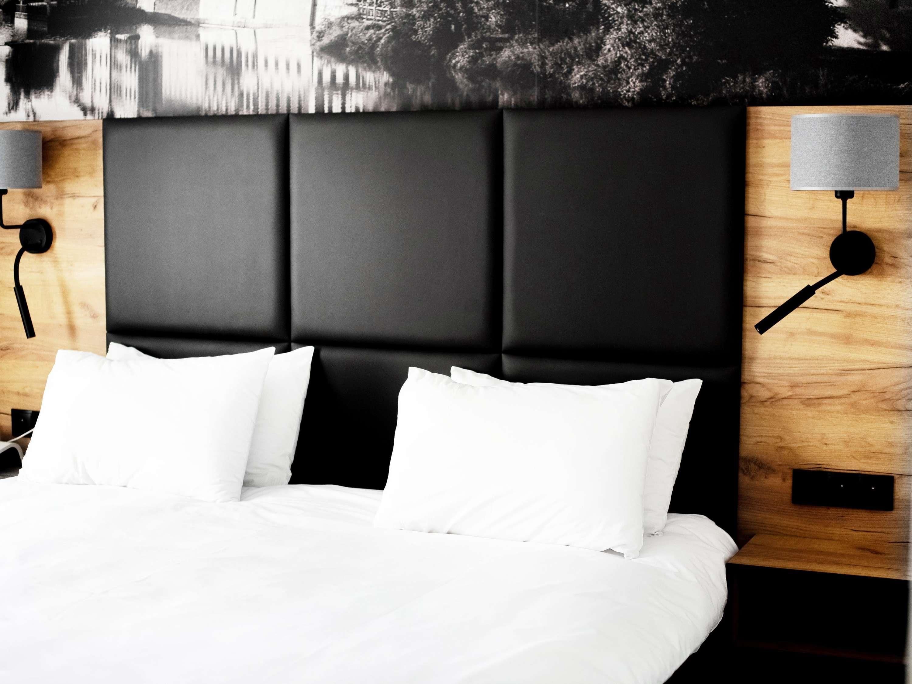 A modern hotel room with a large bed, black headboard, white bedding, and two wall-mounted lamps.