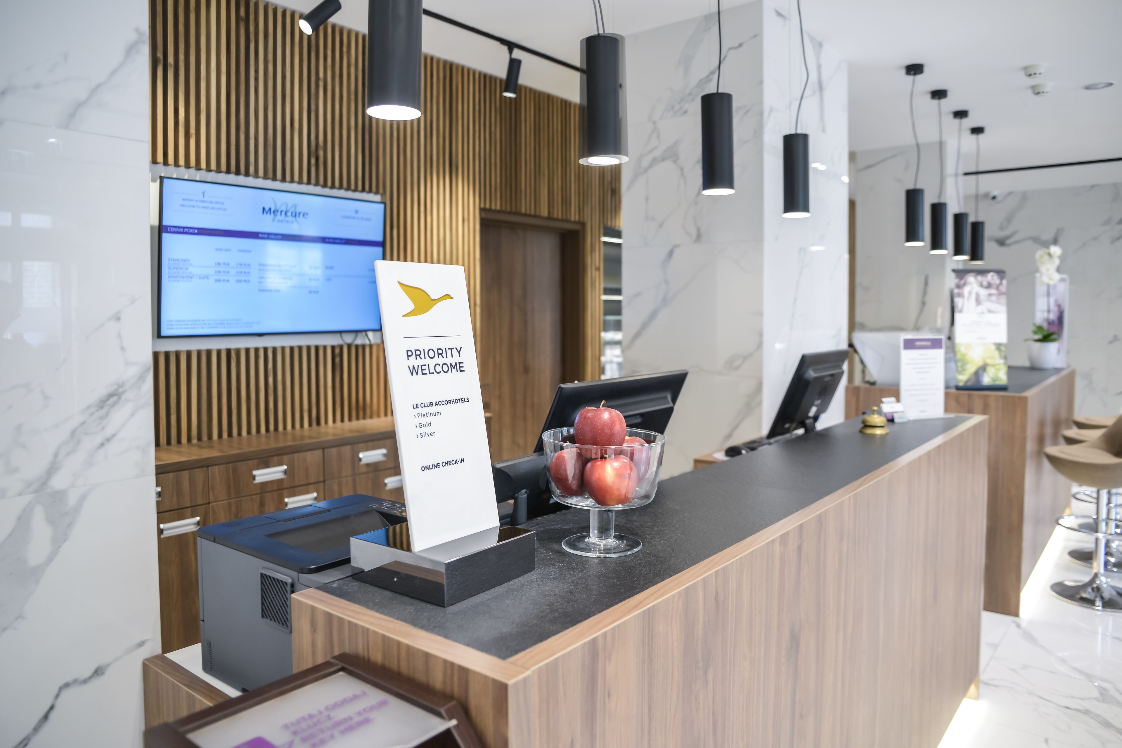 A modern reception desk with a wooden panel backdrop, a digital screen displaying room rates, and a sign indicating priority welcome.