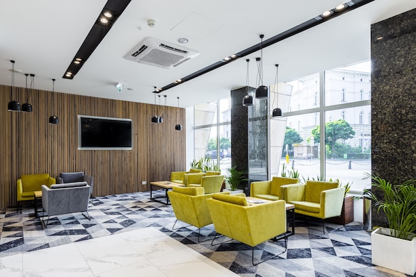 A modern lobby with yellow and gray seating, a large TV, and a check-in counter.