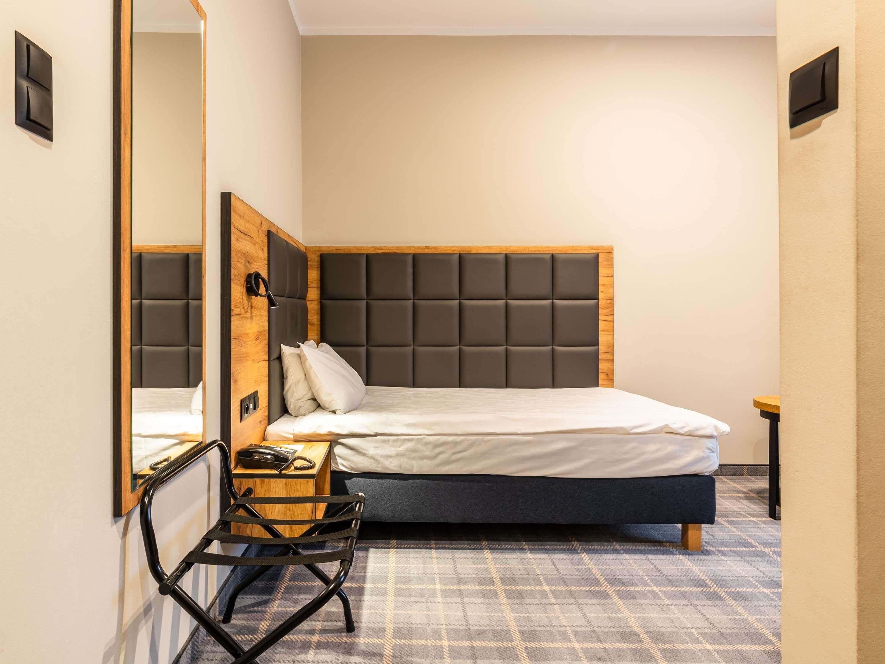 A modern hotel room with a large bed, a wooden headboard, a small bedside table with a lamp, a chair, and a mirror.