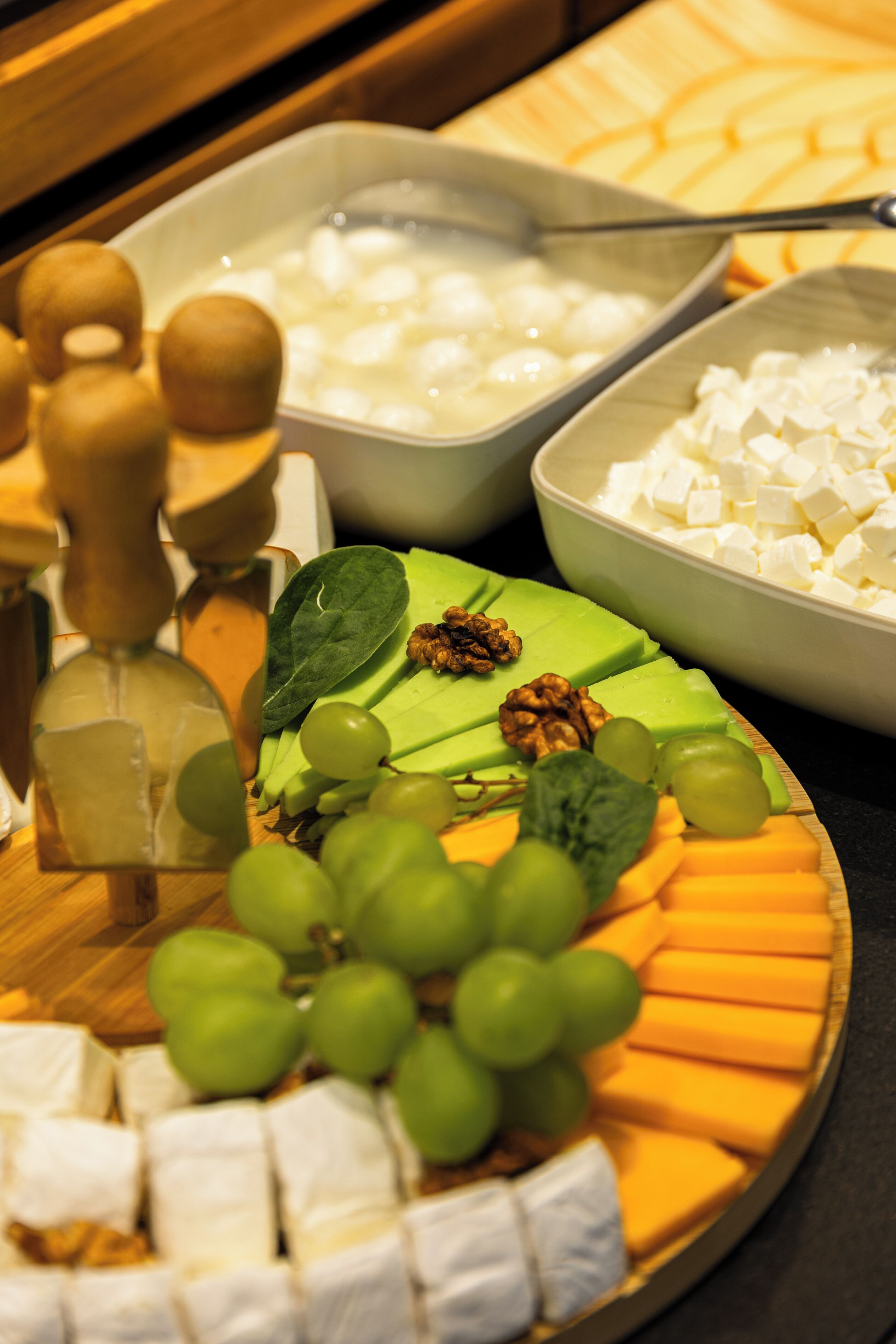 A cheese platter with various cheeses, grapes, walnuts, and leafy greens on a wooden board.