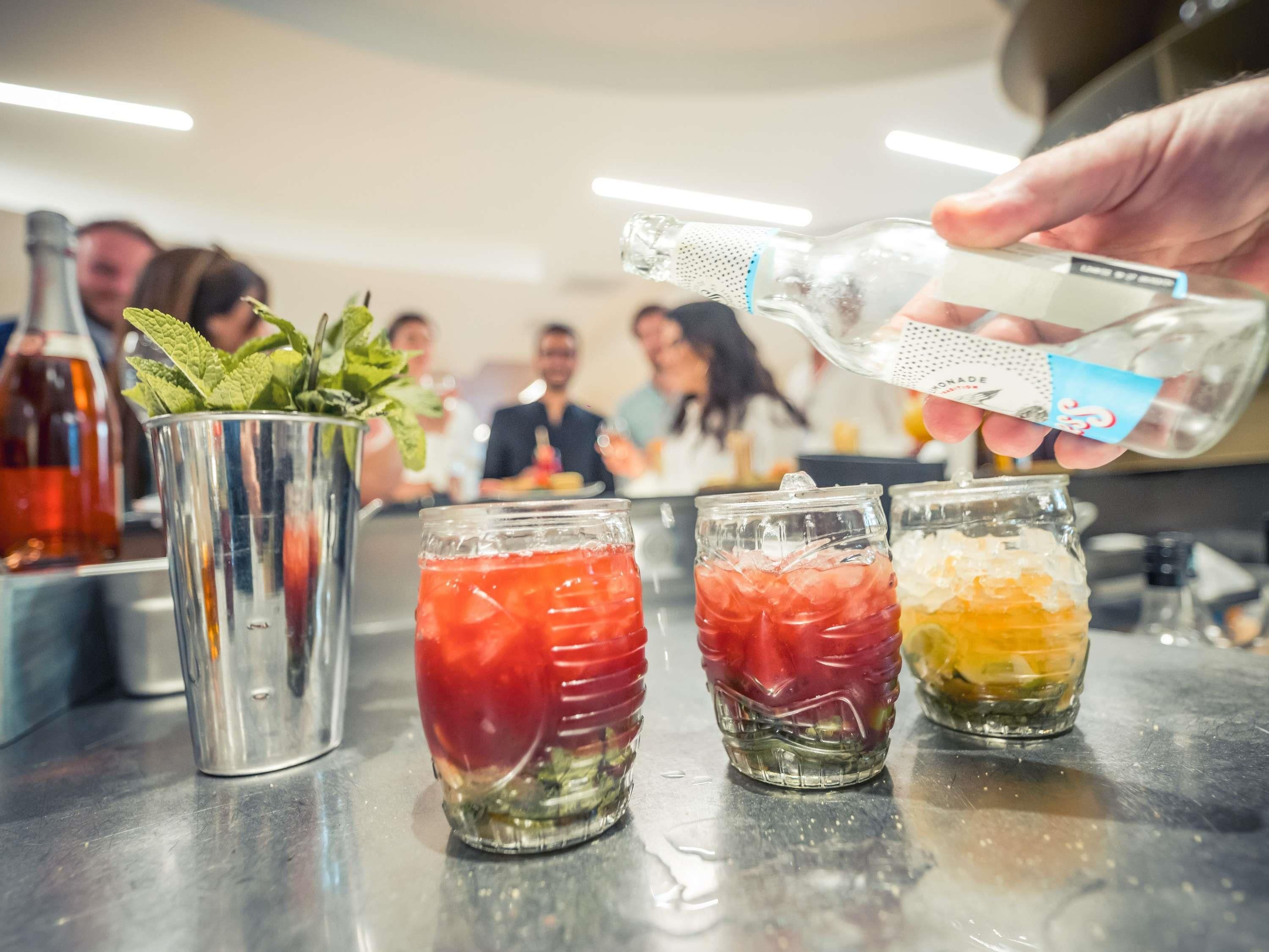 A bar setting with three glasses of mixed drinks, a metal bucket with mint, and a hand pouring a drink.
