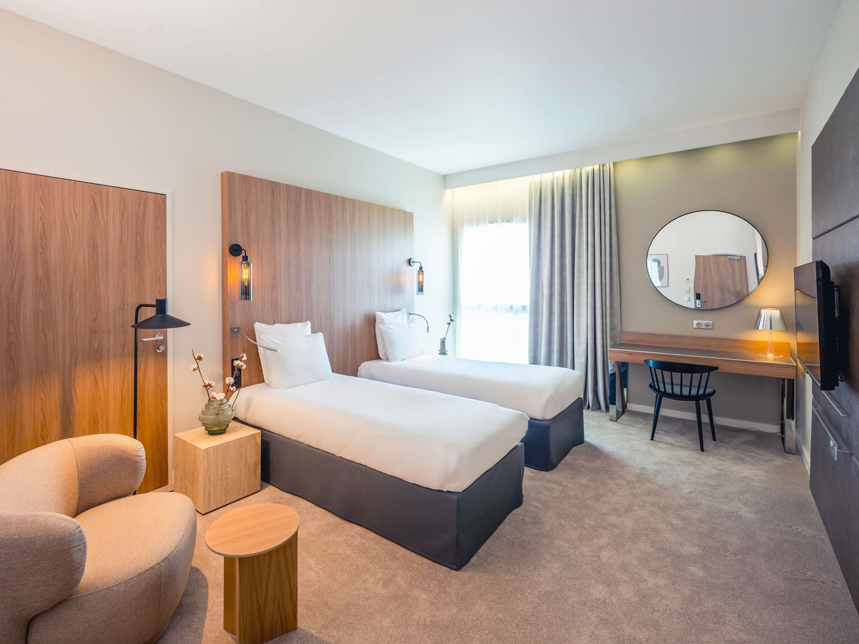 A hotel room with two beds, a desk, a chair, a round mirror, and a TV.