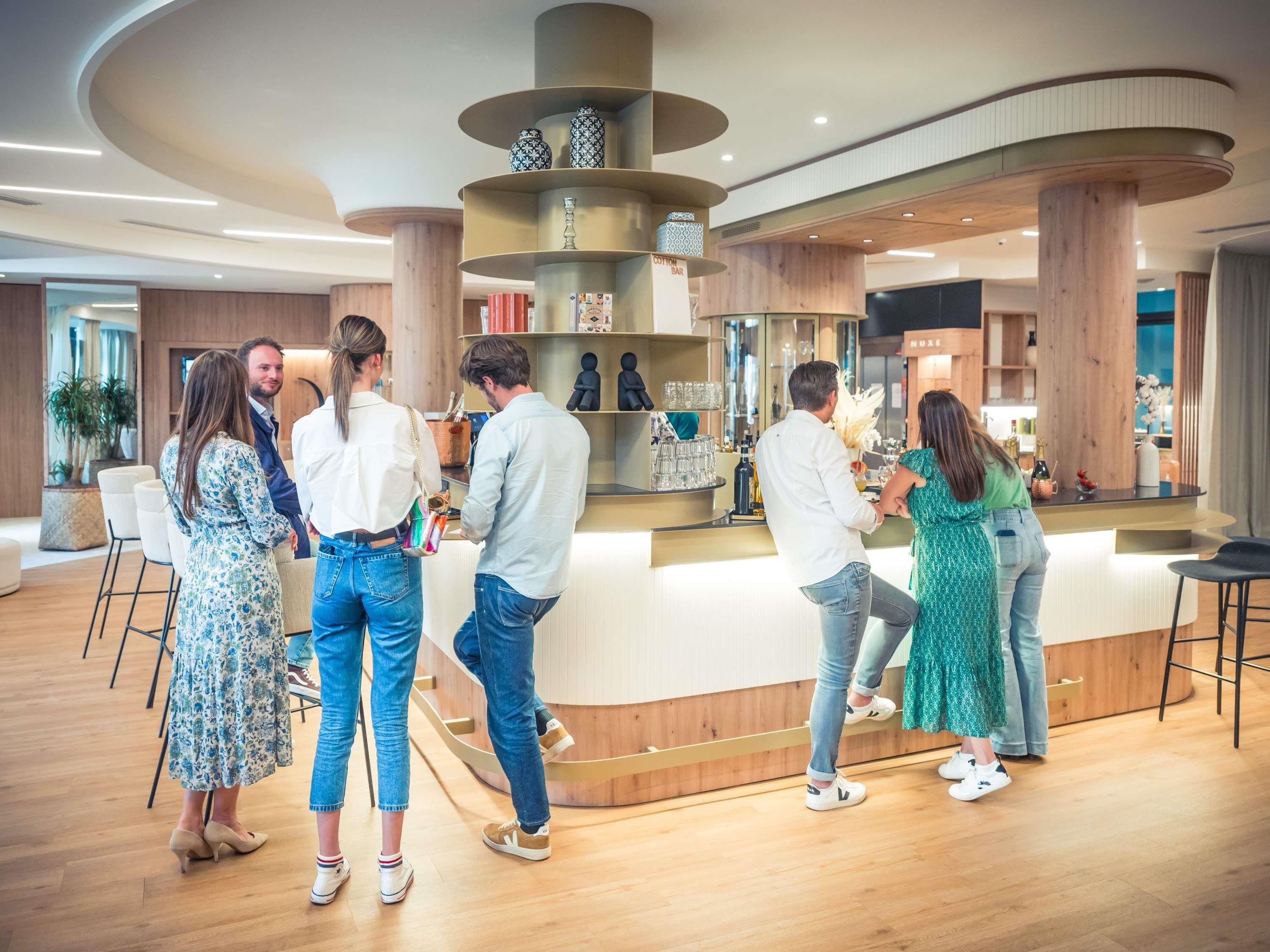 Un moderno vestíbulo de hotel con una zona de bar central, asientos distribuidos y un mostrador de recepción de madera.