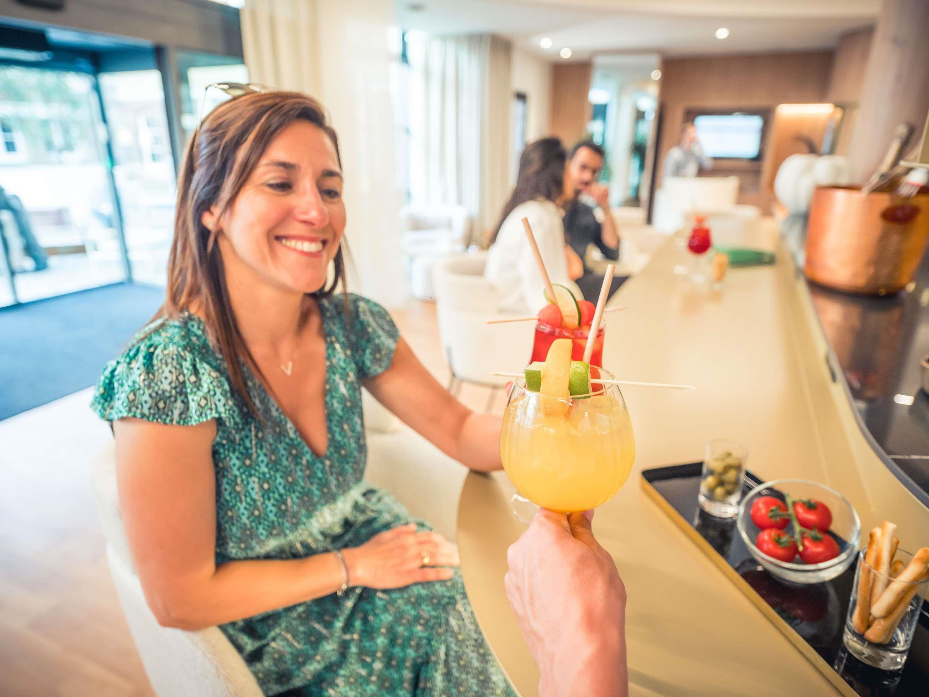 Une personne tenant un cocktail décoré d’un fruit tropical, assise dans le hall d’un hôtel moderne, qui comprend un bar et un espace salon.