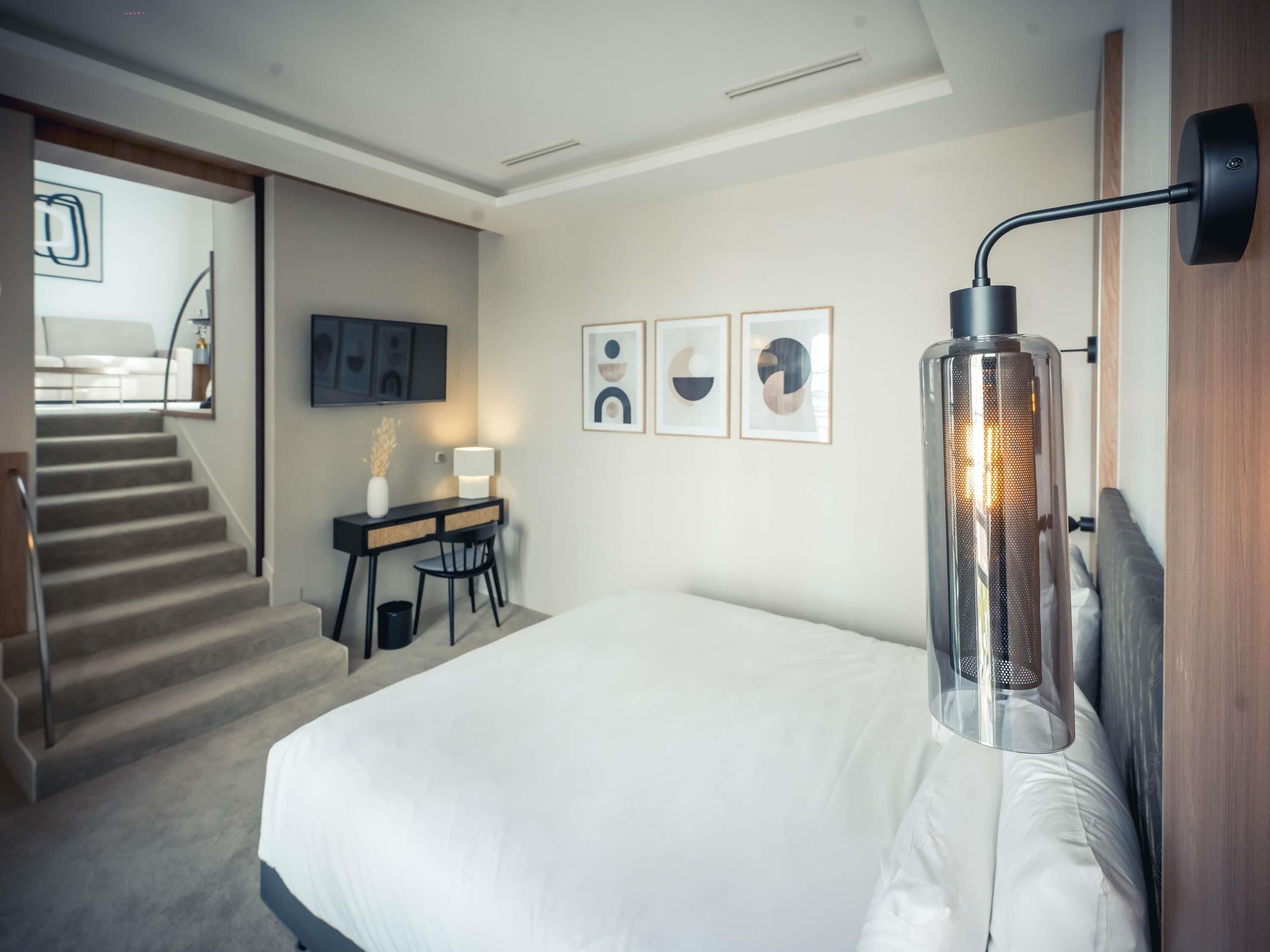 A modern hotel room with a bed, desk, chair, and staircase.
