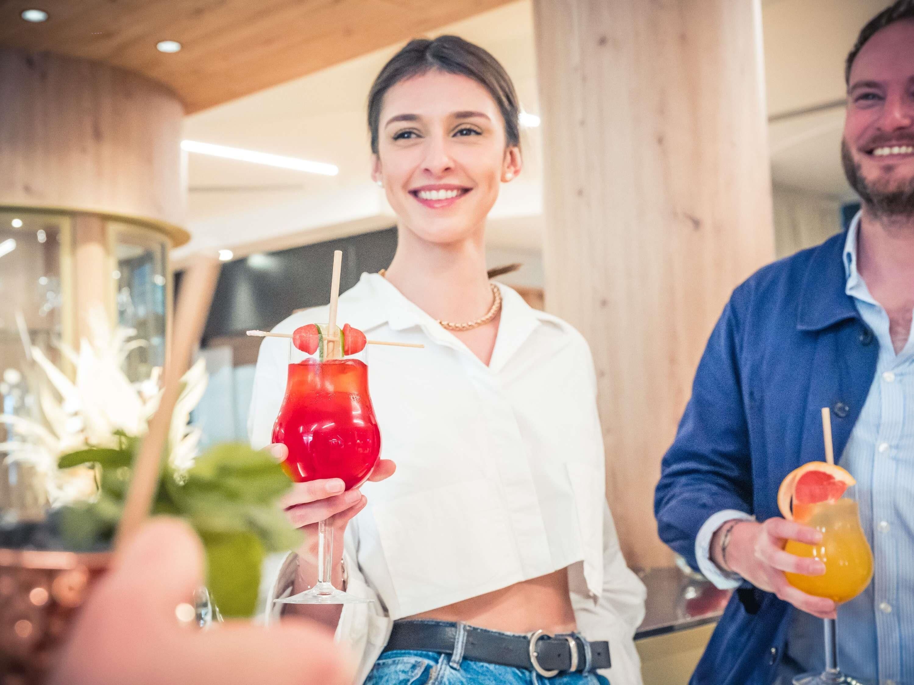 Yksi henkilö pitää kädessään punaista cocktail-lasia, jossa on koriste, toinen pitää kädessään cocktail-lasia, jossa on sitruunaviipale, ja kolmas henkilö on osittain näkyvissä.