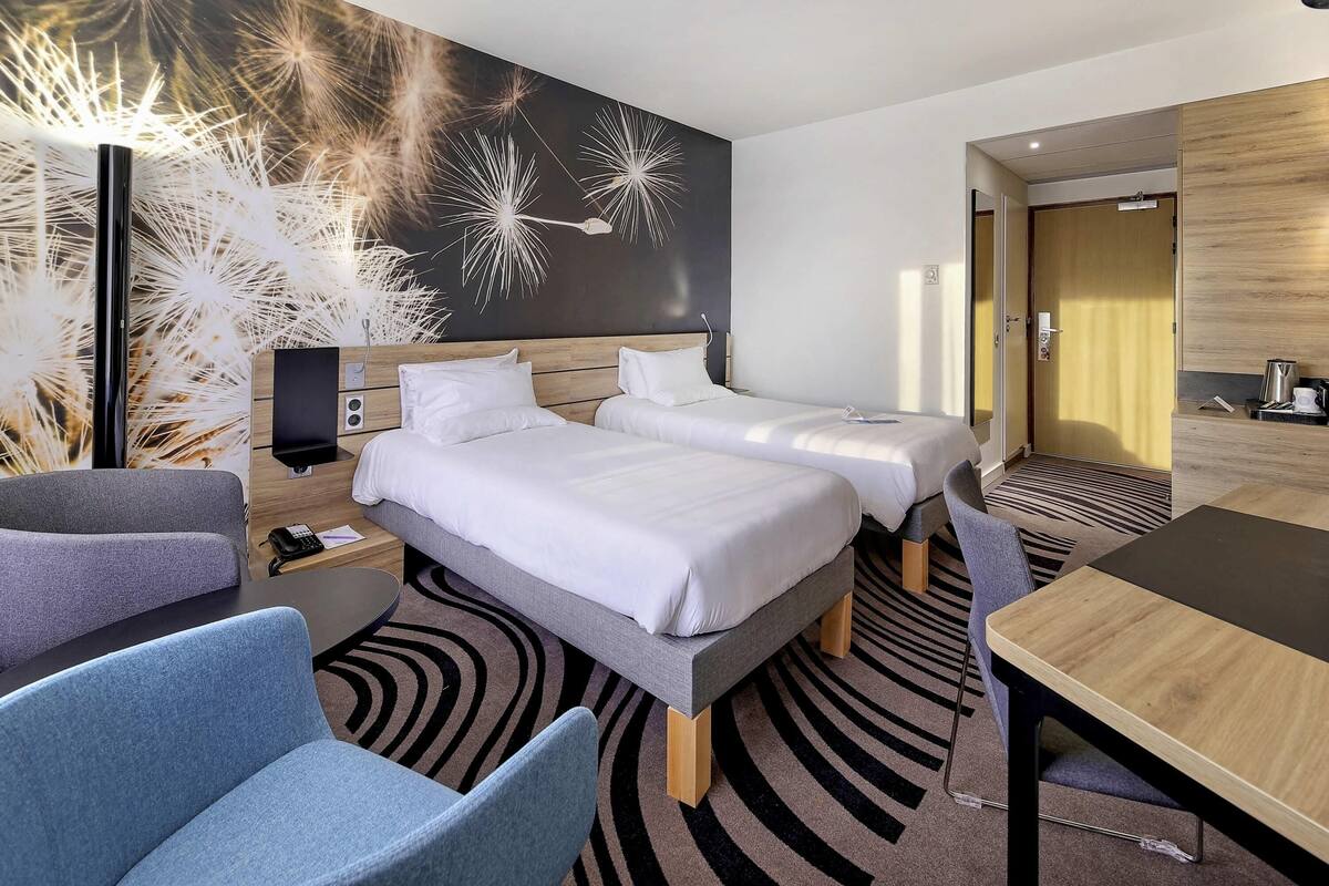 A hotel room with two beds, a desk, a chair, and a wall with a dandelion pattern.