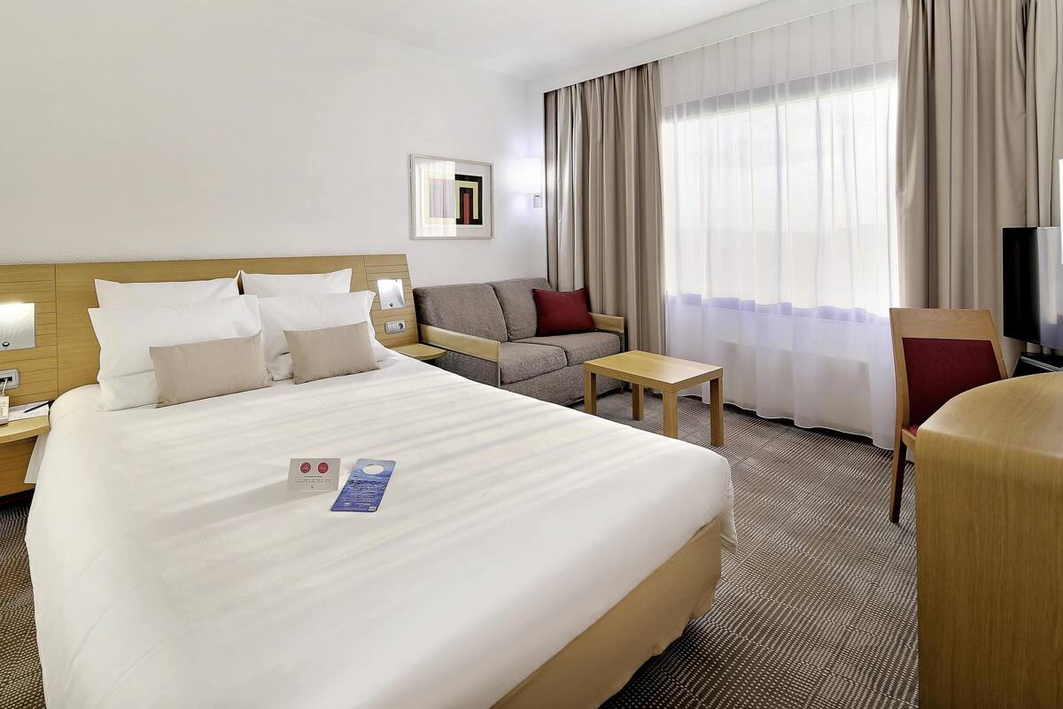 A hotel room with a large bed, a sofa, a small table, a TV, and a window with curtains.
