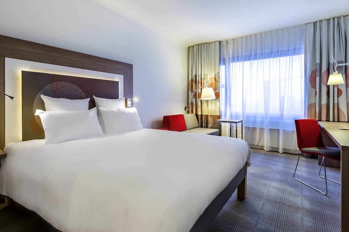 A hotel room with a large bed, a desk, a chair, and a window with curtains.
