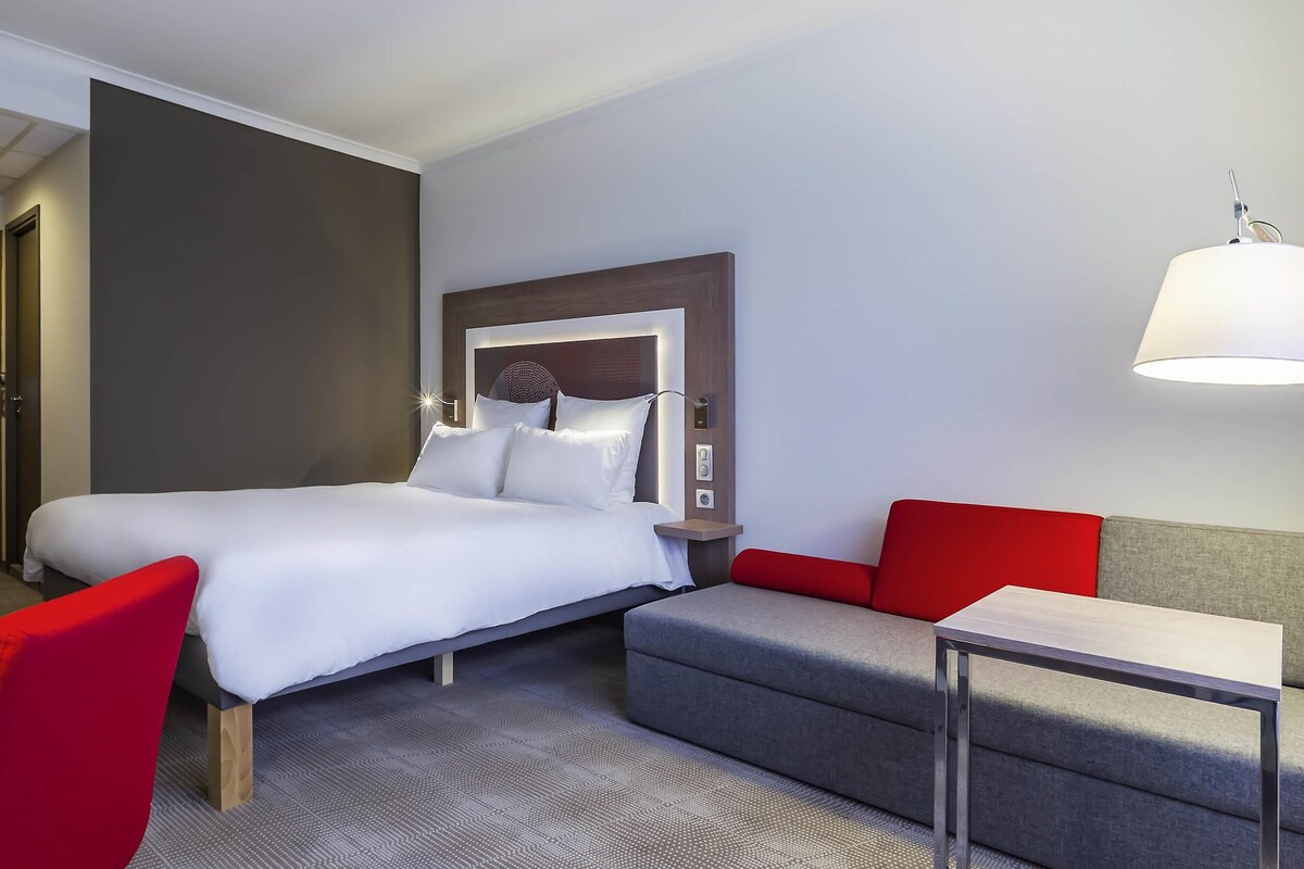 A hotel room with a bed, a desk, a red chair, a grey sofa, and a wall-mounted lamp.