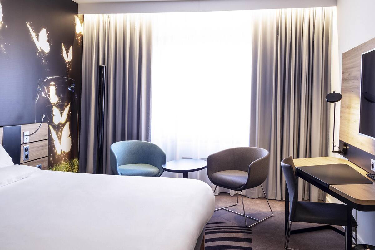 A hotel room with a large bed, two chairs, a small round table, a desk, and a TV.
