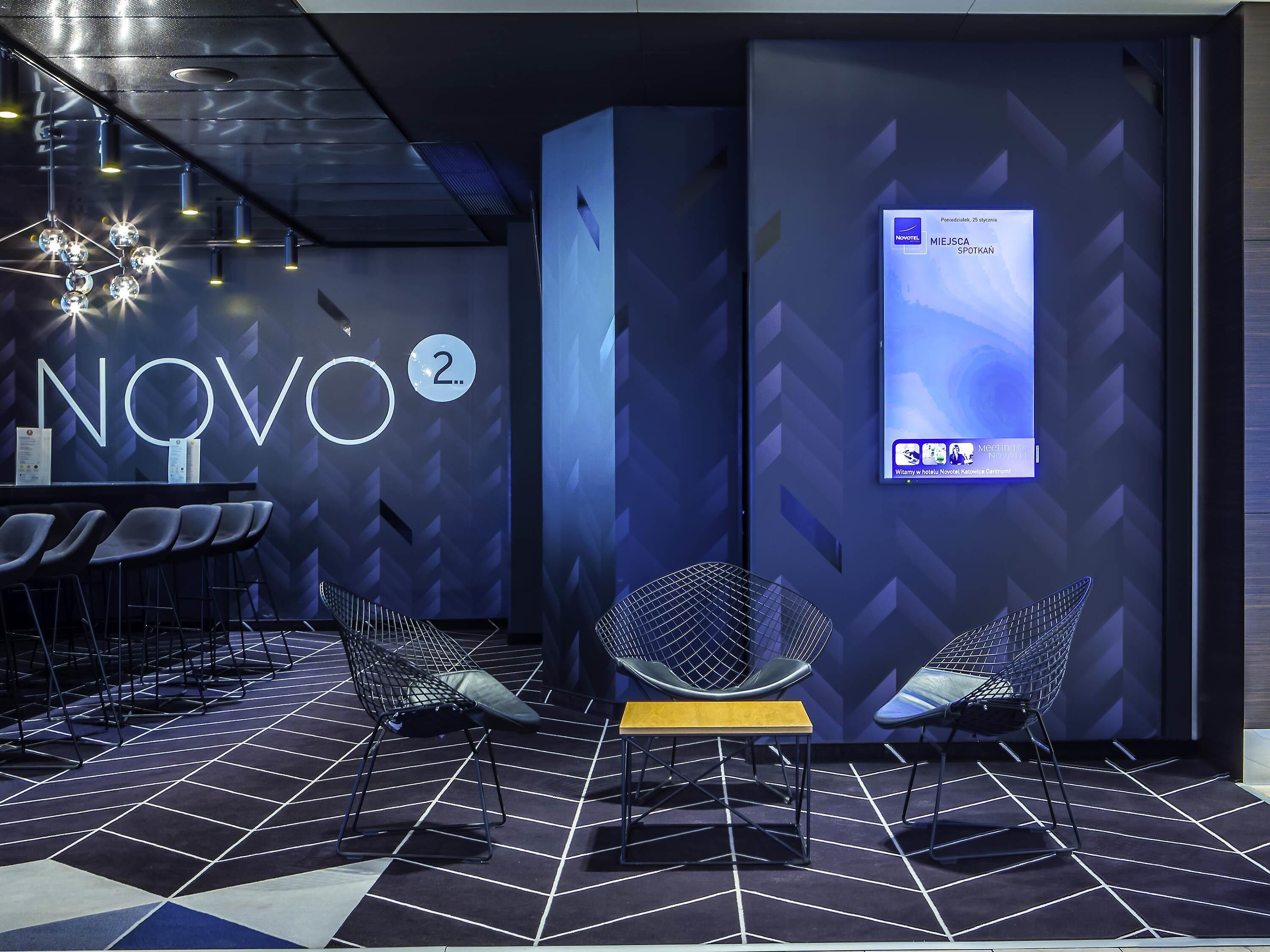 A modern hotel lobby with a geometric floor pattern, a prominent 'NOVO 2' sign, and a digital display on the wall.