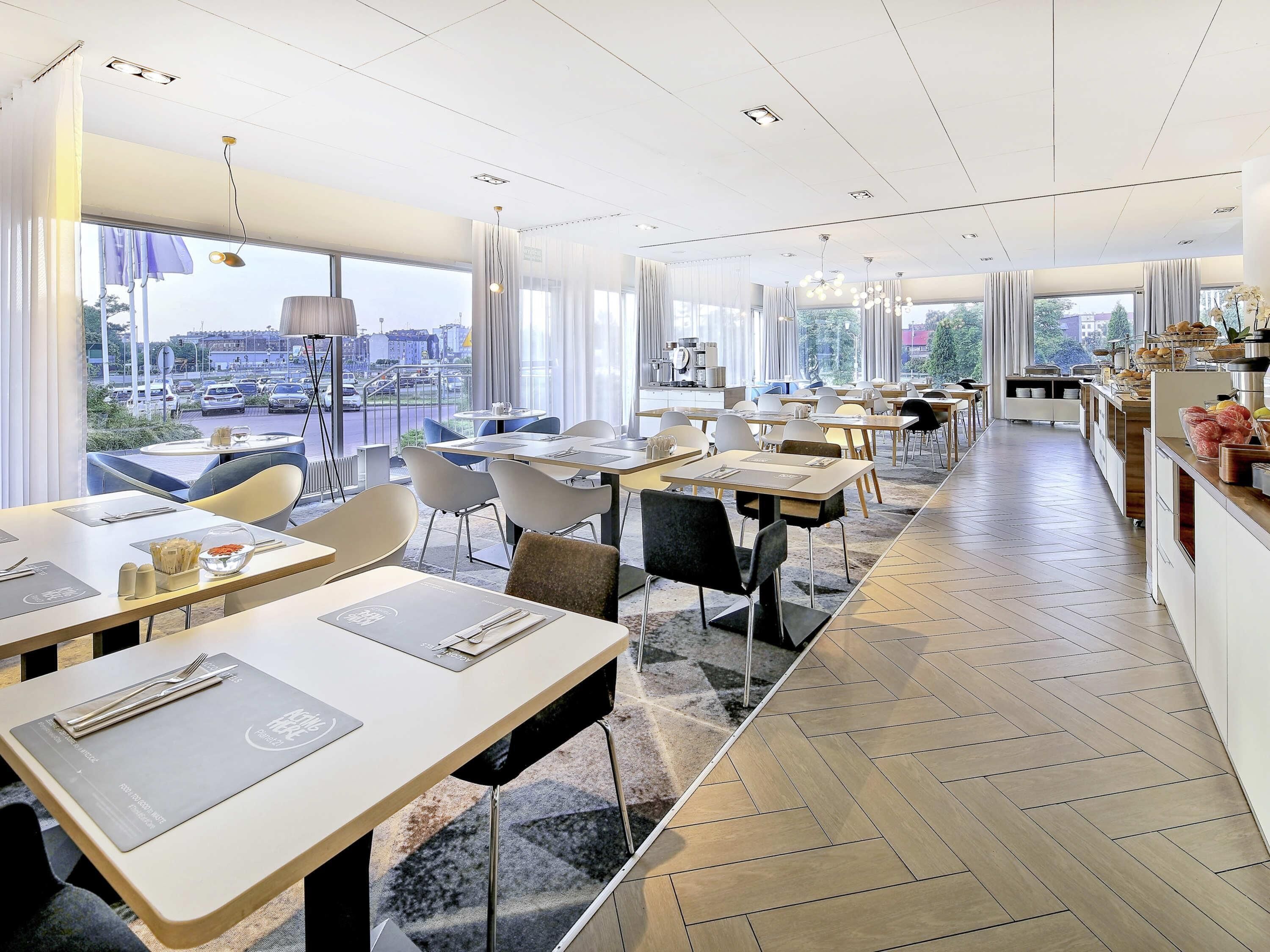 Un restaurante moderno con varias mesas y sillas, grandes ventanales que ofrecen vistas a la ciudad, y un diseño interior limpio y contemporáneo.