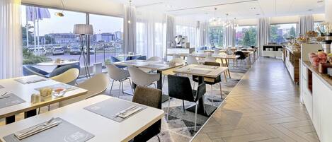 Un restaurante moderno con varias mesas y sillas, grandes ventanales que ofrecen vistas a la ciudad, y un diseño interior limpio y contemporáneo.