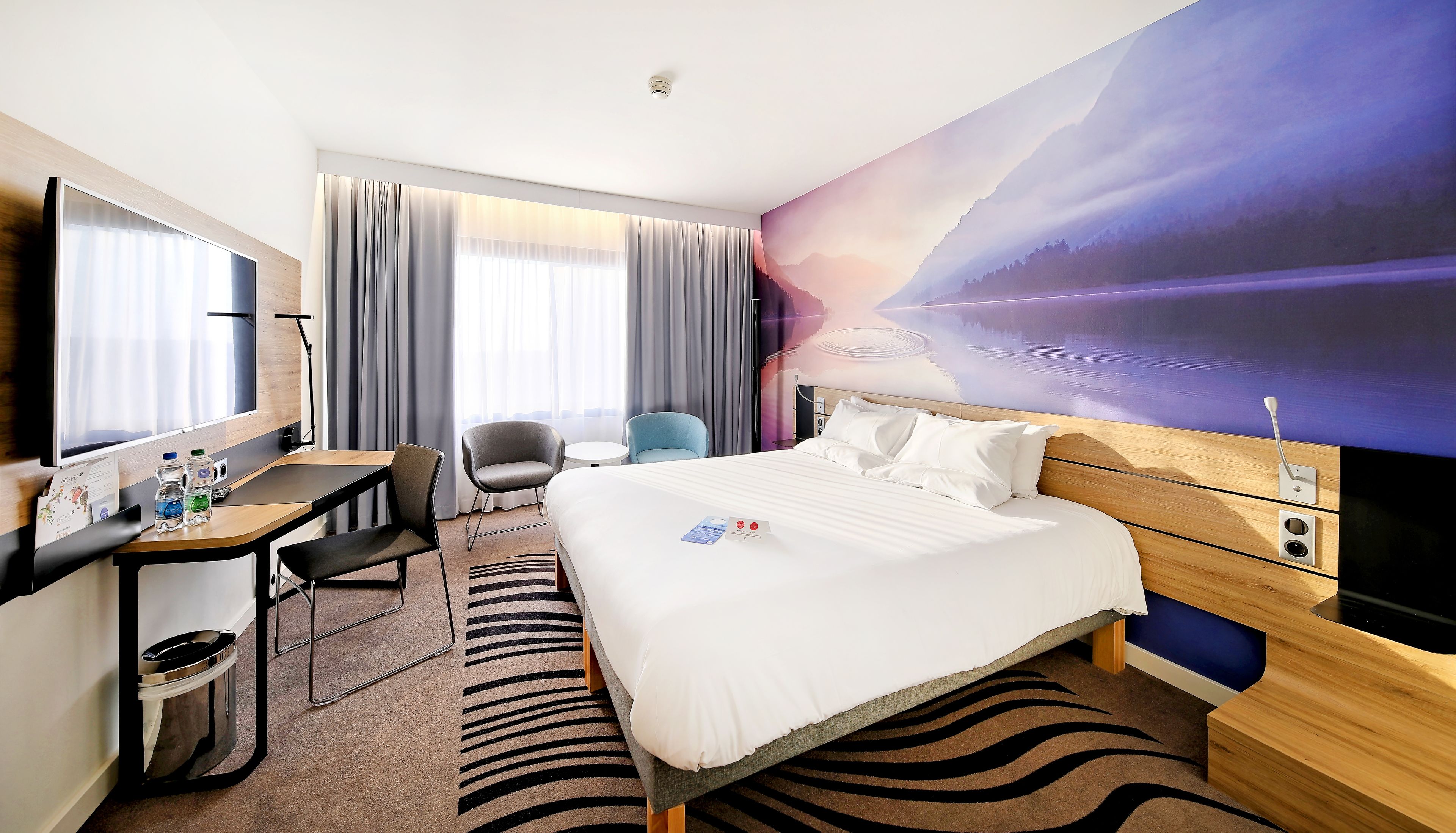 A hotel room with a bed, a desk, a chair, a TV, and a large window with curtains.