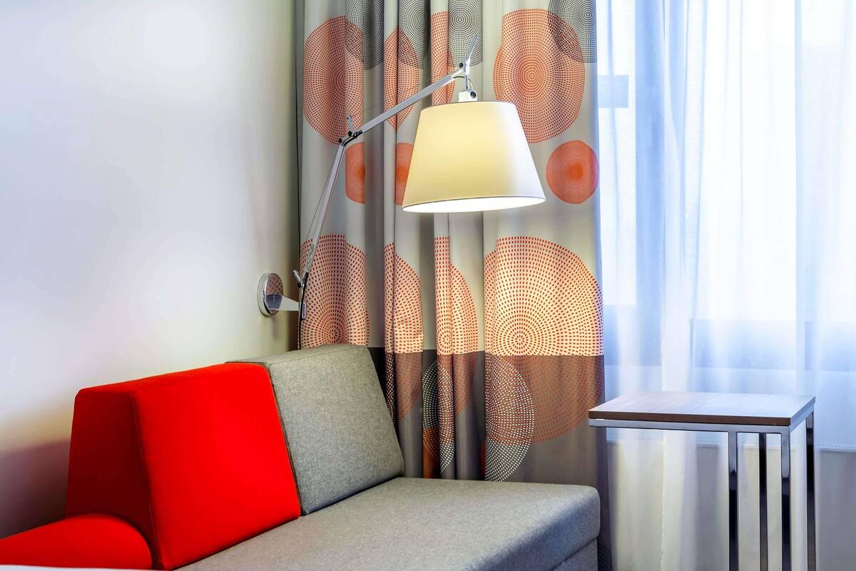 A modern hotel room with a red and grey sofa, a table, and a lamp.
